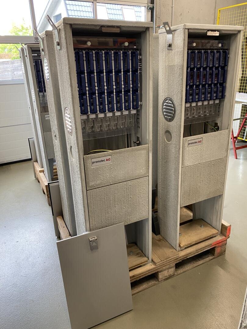 Zwei große graue Schaltschränke mit offenen Fronten, die den Blick auf blaue Bauteile und Kabel im Inneren freigeben, stehen auf Holzpaletten in einer Industriehalle.