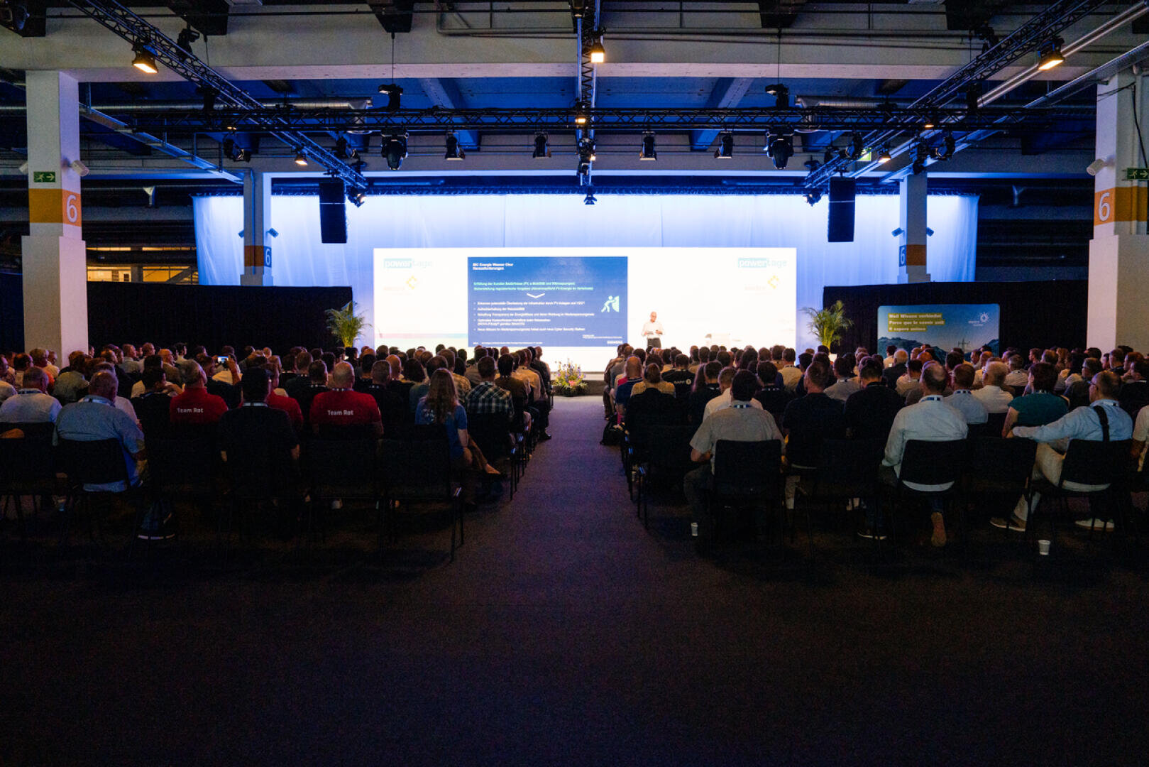 Ein großes Publikum sitzt in einem Konferenzsaal und blickt auf eine hell beleuchtete Bühne, auf der ein Redner vor einer großen Leinwand, auf der Folien gezeigt werden, seinen Vortrag hält.