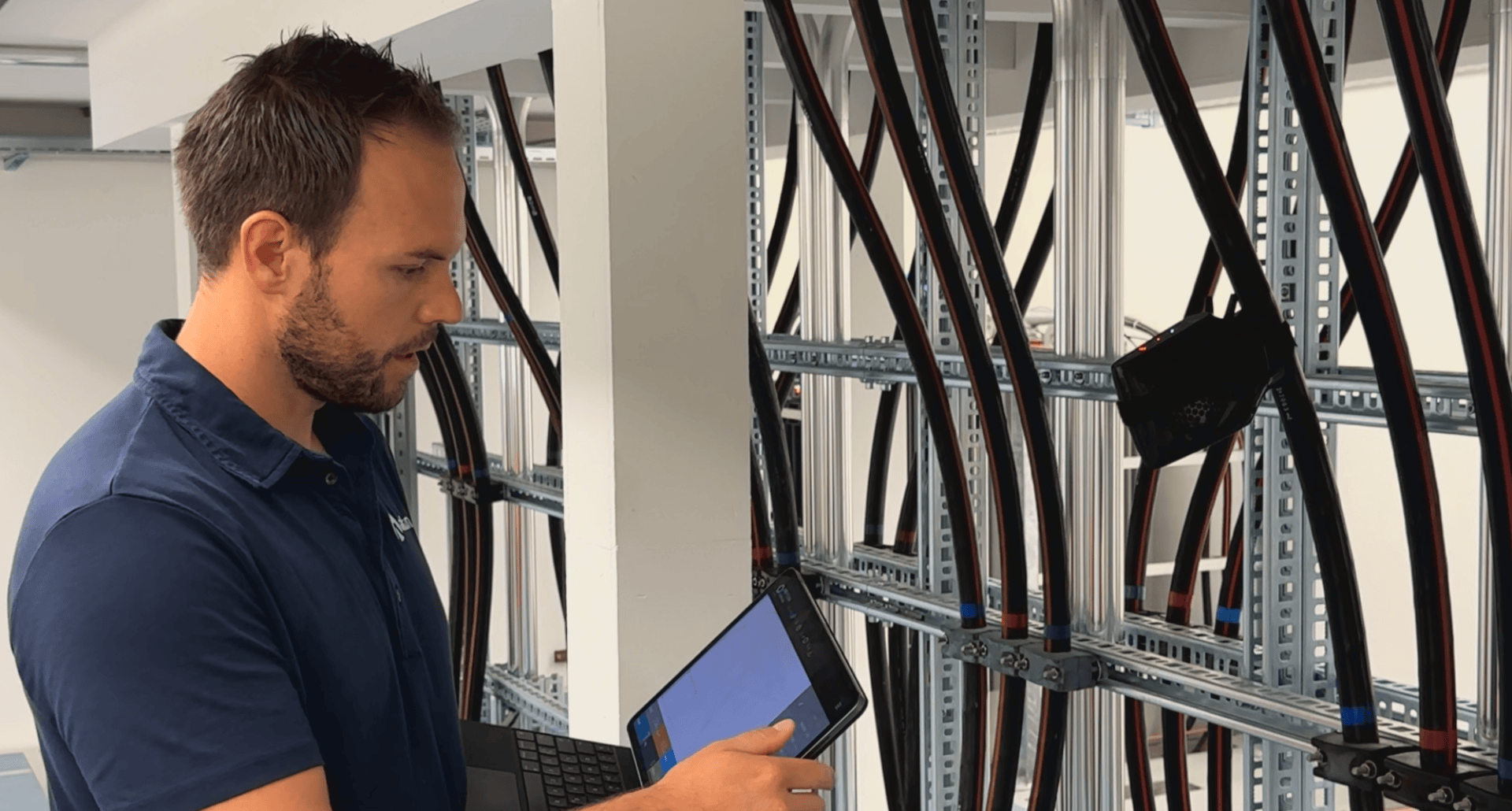 Ein Mann in einem blauen Hemd prüft große elektrische Kabel an Regalen, hält ein Tablet und einen Laptop in der Hand und scheint Daten zu prüfen oder Wartungsarbeiten in einer industriellen Umgebung durchzuführen.