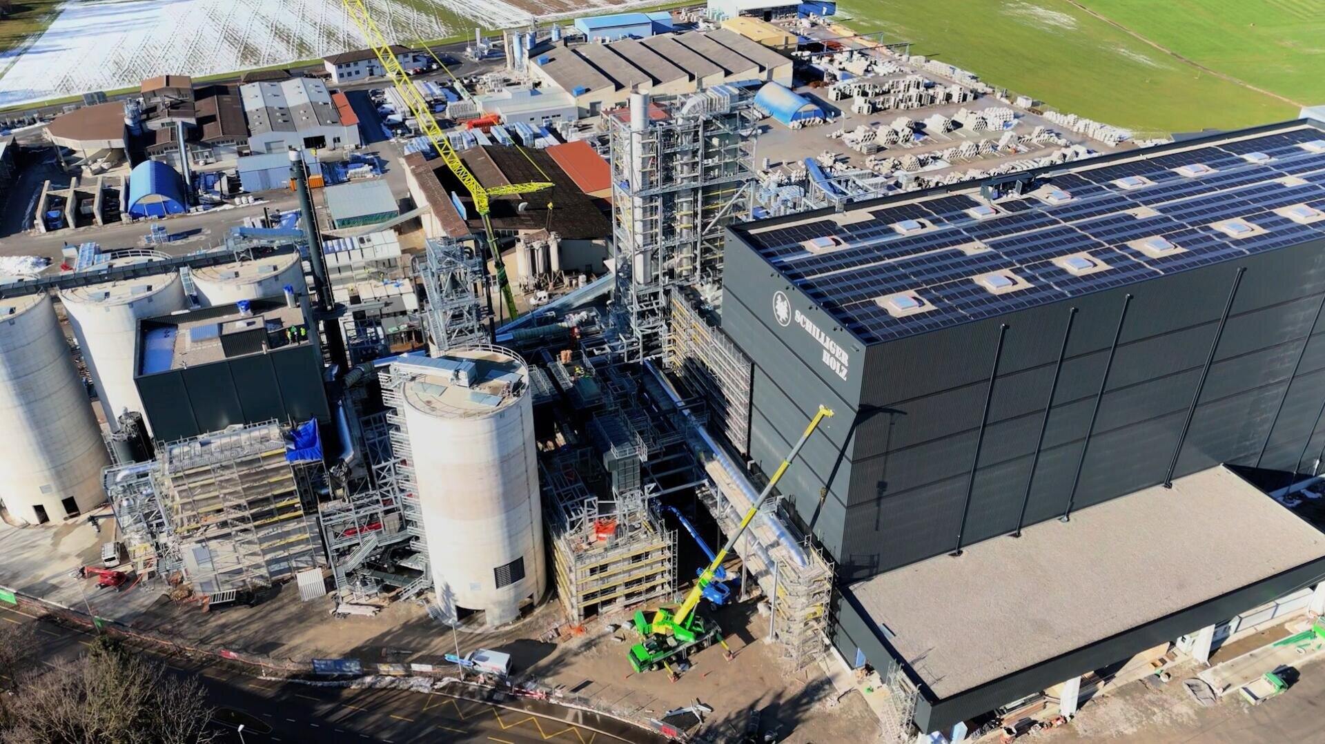 Luftaufnahme einer Industrieanlage mit großen Tanks, Gerüsten und einem schwarzen Gebäude mit Solarzellen auf dem Dach; am Boden sind Baufahrzeuge und Arbeiter zu sehen.