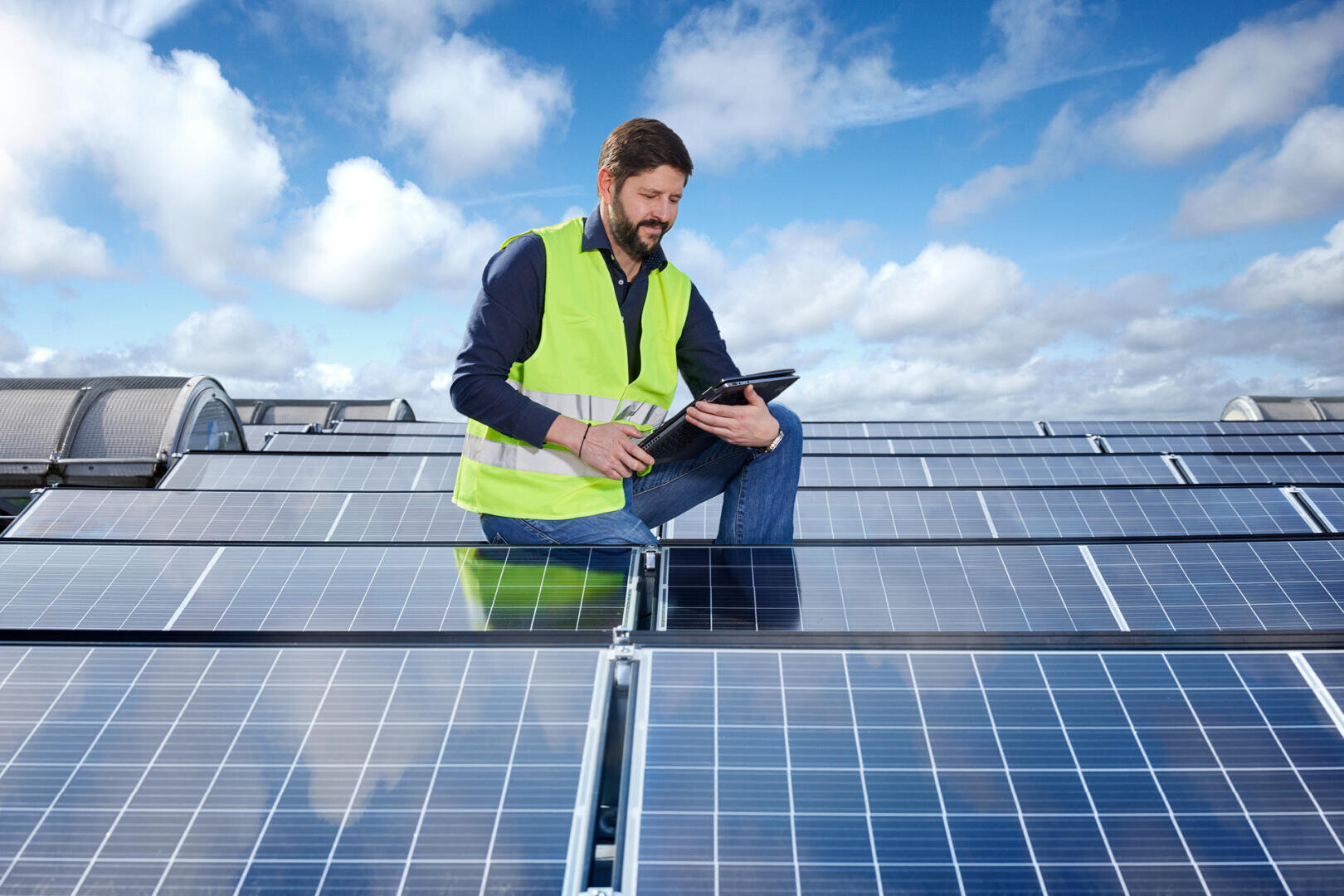Ein Mann in einer gelben Sicherheitsweste kniet auf einem Dach zwischen Solarmodulen, hält ein Tablet in der Hand und inspiziert die Module unter einem teilweise bewölkten Himmel.