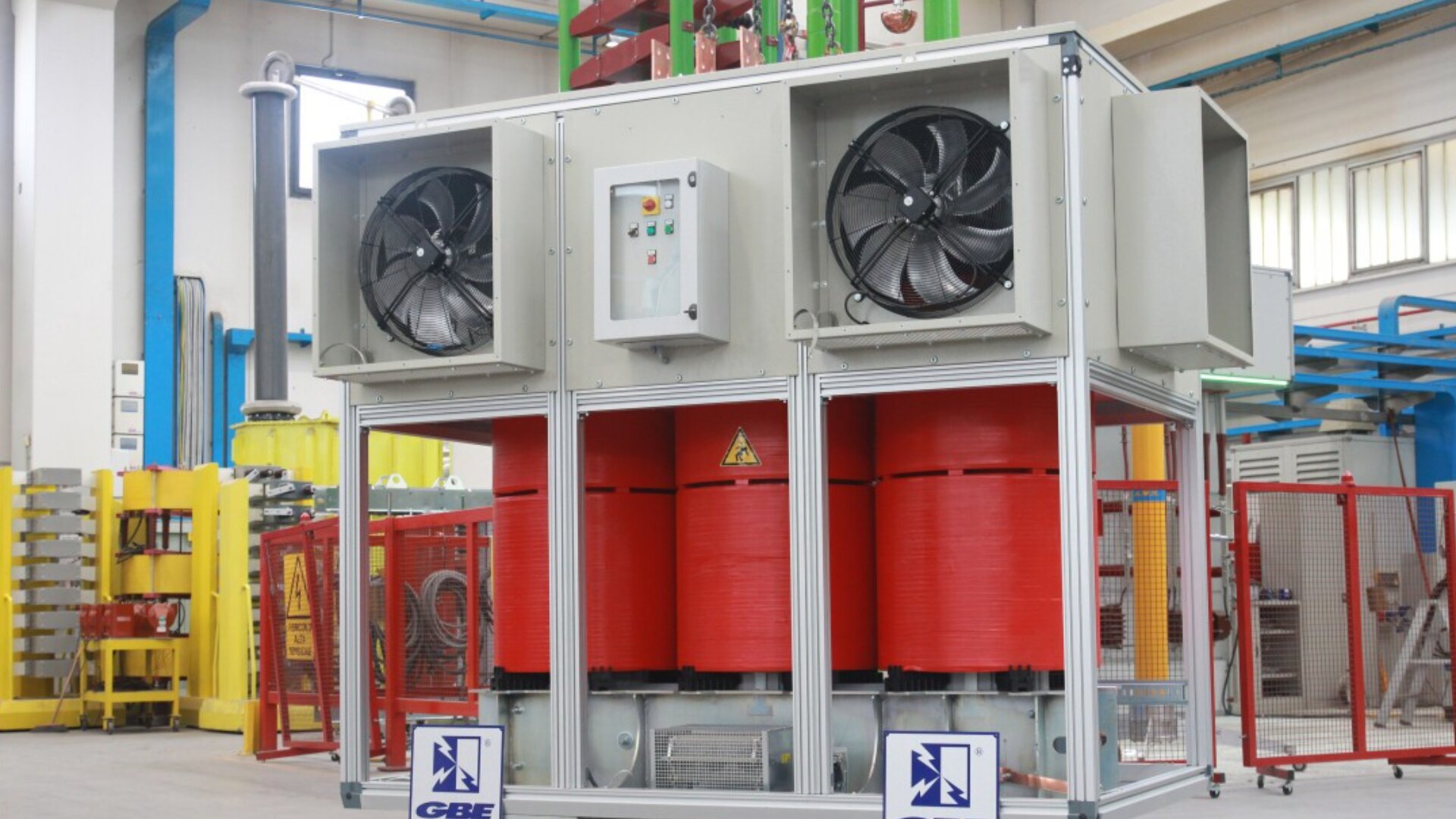 Ein großer elektrischer Industrietransformator mit roten zylindrischen Spulen und zwei schwarzen Kühlgebläsen befindet sich in einer Werkstatt mit blauen Balken und Maschinen im Hintergrund. Auf dem Transformator sind Sicherheits- und Herstelleretiketten zu sehen.