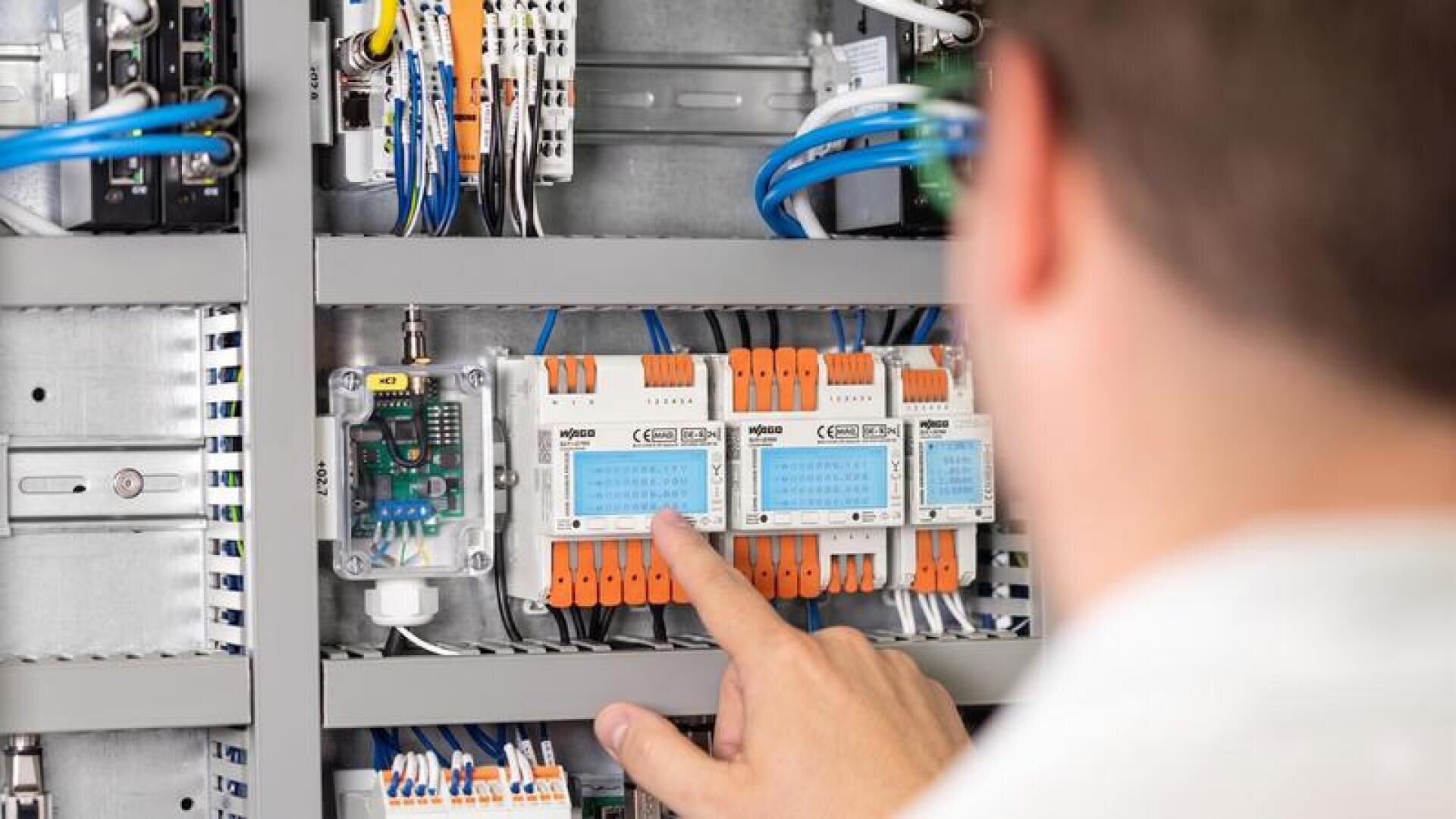 Person vor einem geöffneten Schaltschrank mit mehreren installierten Energiezählern und elektrischen Komponenten. Im Vordergrund zeigt eine Hand auf ein Gerät im Schaltschrank. Die Aufnahme vermittelt eine typische Anwendungssituation für Integration und Konfiguration von Messgeräten in einer technischen Anlage.