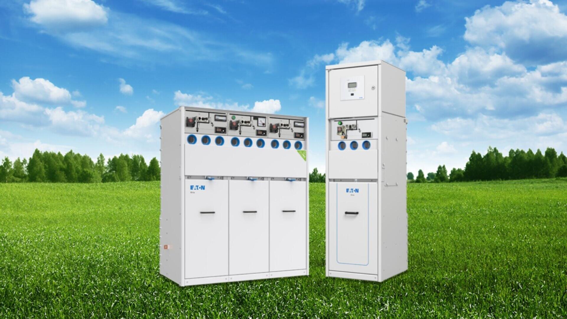 Two white electrical switchgear cabinets with control panels and blue labels stand on a grassy field, with a backdrop of trees and a bright blue sky with scattered clouds.