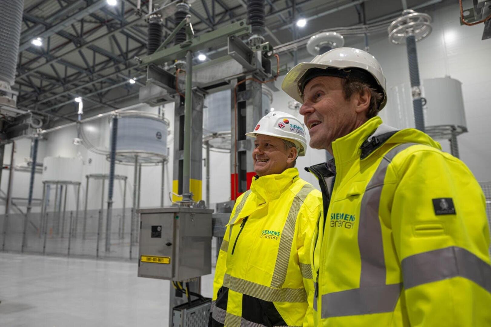 Zwei Personen in gelben Siemens-Energy-Jacken und weißen Schutzhelmen stehen in einer Industrieanlage mit großen Maschinen und Metallkonstruktionen und scheinen den Standort zu begutachten oder zu besprechen.
