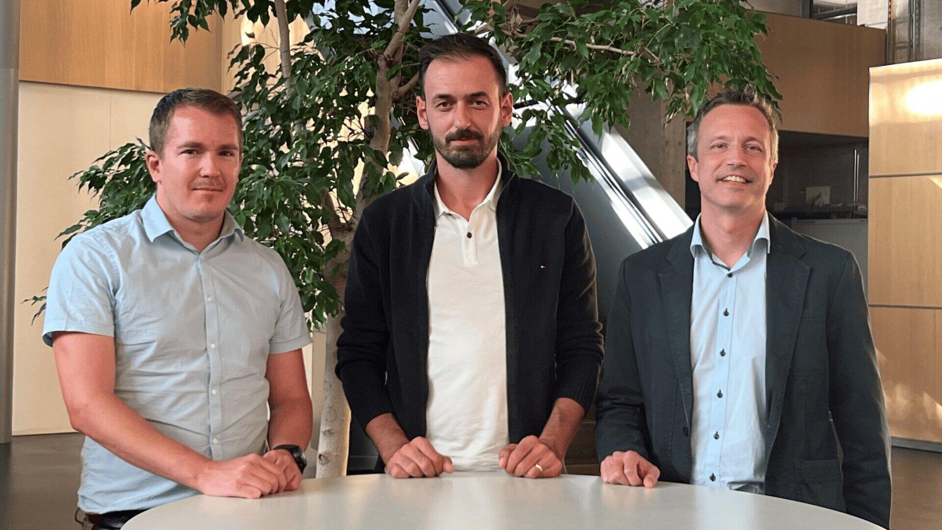 Drei Männer stehen Seite an Seite hinter einem runden weißen Tisch in einem Innenraum, mit einer grünen Pflanze und modernen Büroelementen im Hintergrund. Sie sind alle leger gekleidet und schauen leicht lächelnd in die Kamera.