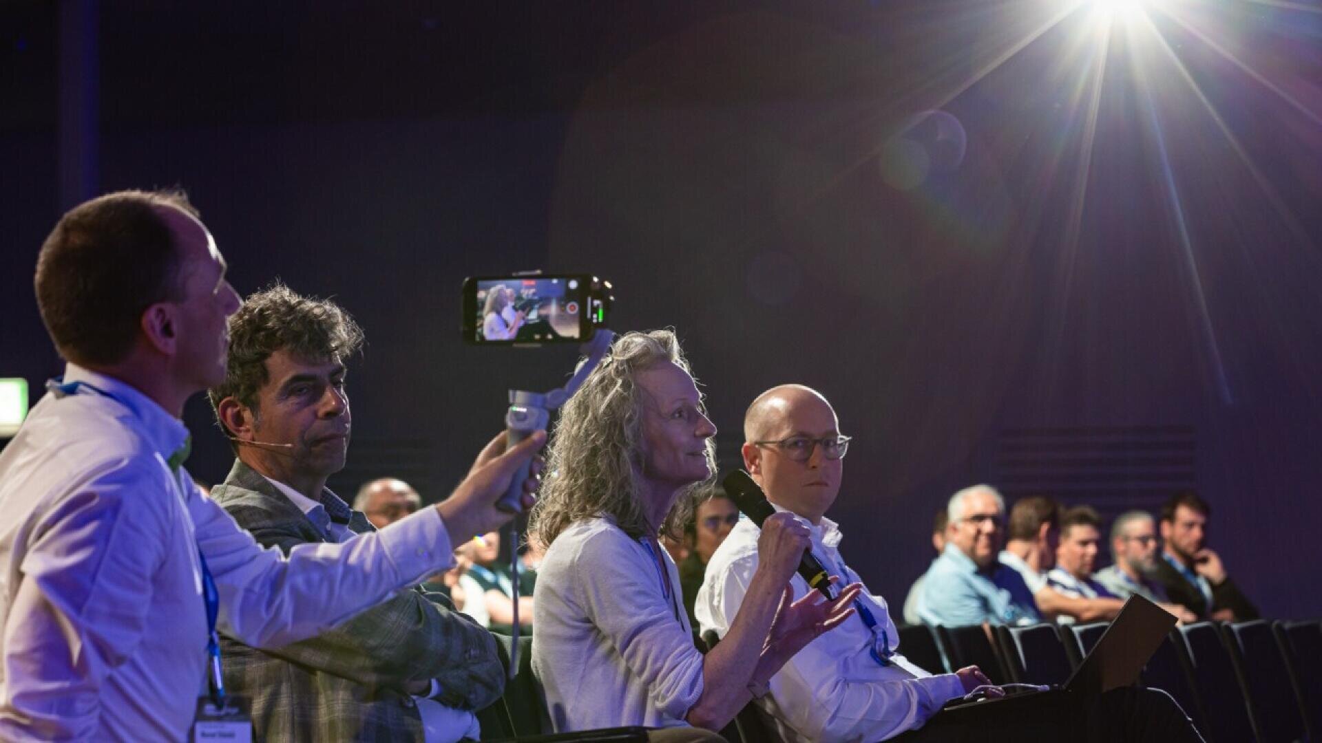 A woman speaks into a microphone at a conference, surrounded by seated attendees. A man beside her films with a smartphone, while others listen attentively under bright stage lighting.