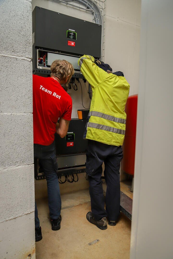 Zwei Personen arbeiten in einem kleinen Hauswirtschaftsraum an elektrischen Geräten. Einer trägt ein rotes Team-Rot-Hemd, der andere eine gelbe Warnweste. Beide stehen mit dem Gesicht zu den Geräten und konzentrieren sich auf die Verkabelung oder die Steuerung.