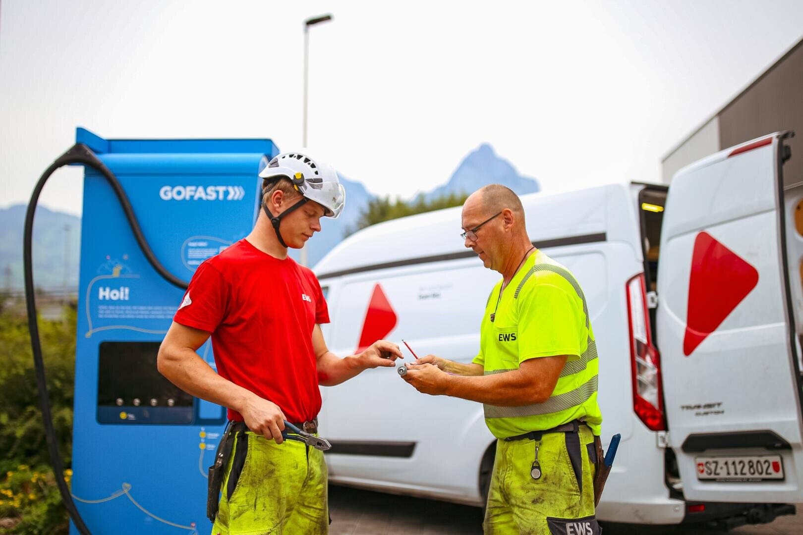Zwei Mitarbeiter eines Energieversorgers, einer mit rotem Hemd und Helm, der andere mit gelbem Hemd, stehen neben einer GOFAST-Ladestation und weißen Servicewagen und diskutieren über Papierkram oder Ausweise. Im Hintergrund sind die Berge zu sehen.