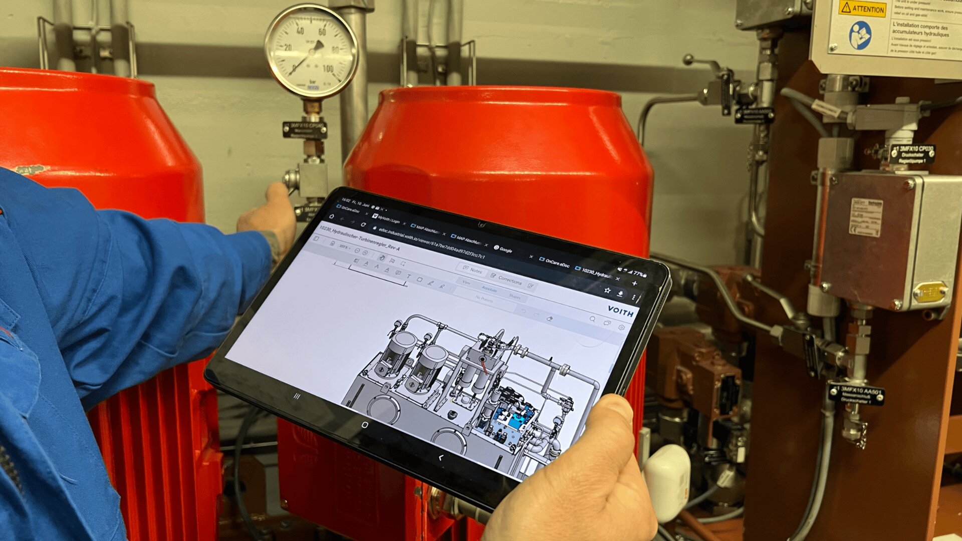 Eine Person in Blau hält ein Tablet in der Hand, auf dem ein technisches Diagramm einer Maschine abgebildet ist, während ihre andere Hand auf ein Druckmessgerät zeigt, das zwischen zwei großen roten Maschinen in einer Industrieumgebung angebracht ist.