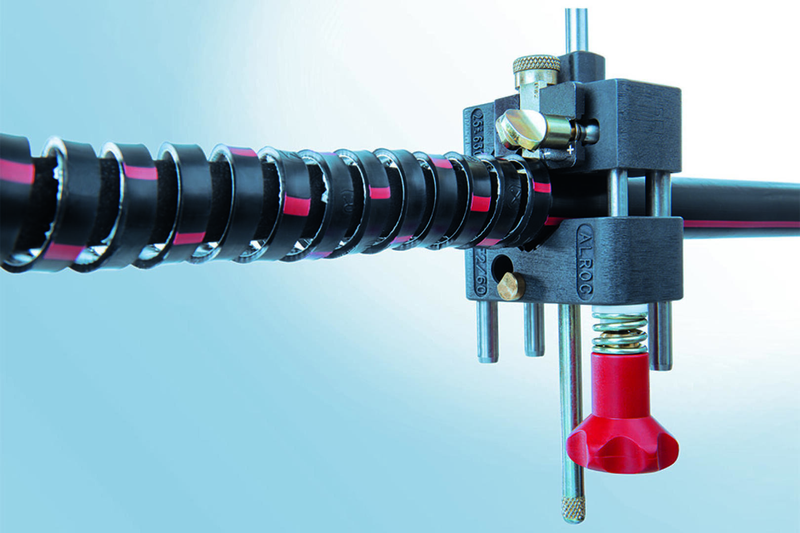 Nahaufnahme eines mechanischen Geräts mit einem schwarzen und roten spiralförmig gewickelten Kabel, Metallstäben, Einstellknöpfen und einem roten Bauteil vor einem blauen Hintergrund. Die Anordnung wirkt technisch oder industriell.