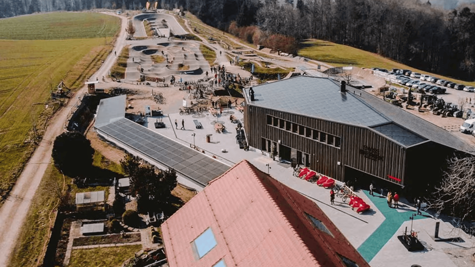 Luftaufnahme eines modernen Fahrradparks mit kurvenreichen Strecken, Rampen und einem großen Holzgebäude; Menschen sind über das Gelände verstreut, und Autos sind am Rand in der Nähe eines bewaldeten Hintergrunds geparkt.