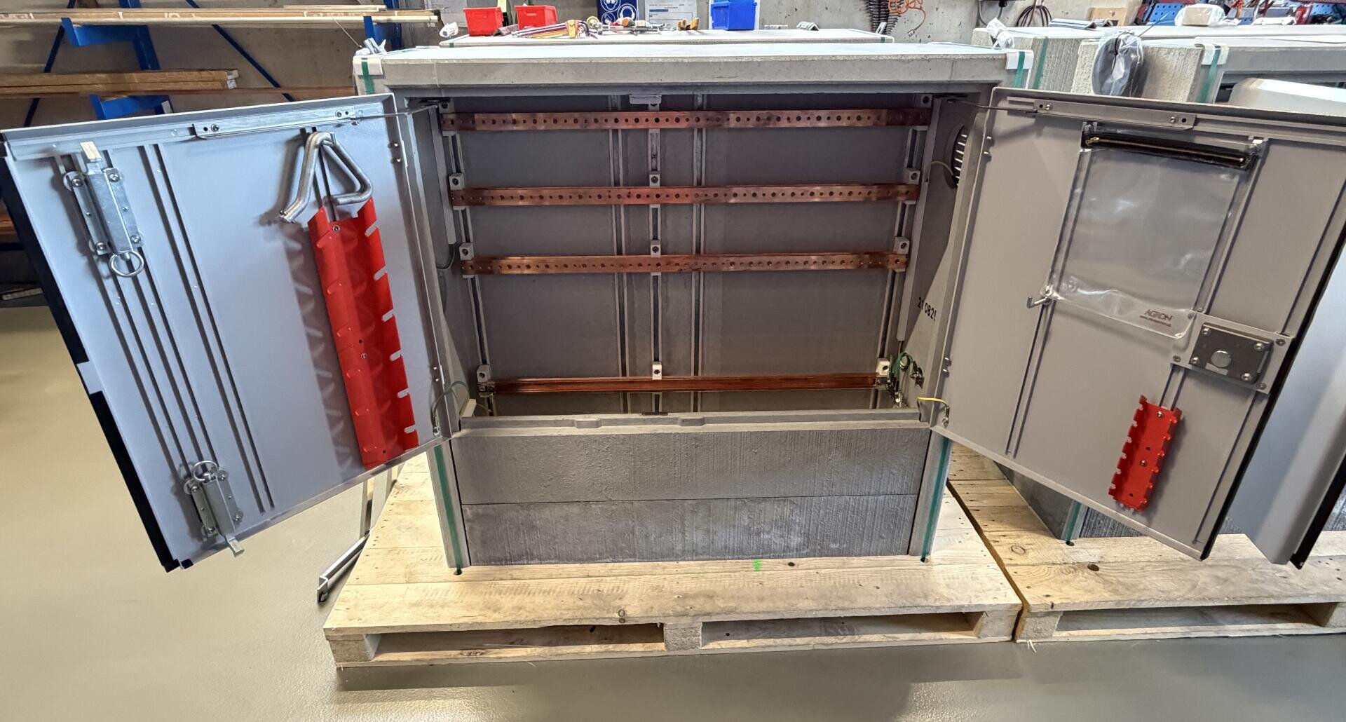 Ein offener industrieller Schaltschrank mit weit geöffneten Türen, in dem leere Kupfersammelschienen und Klemmleisten zu sehen sind, steht auf einer Holzpalette in einer Werkstatt.