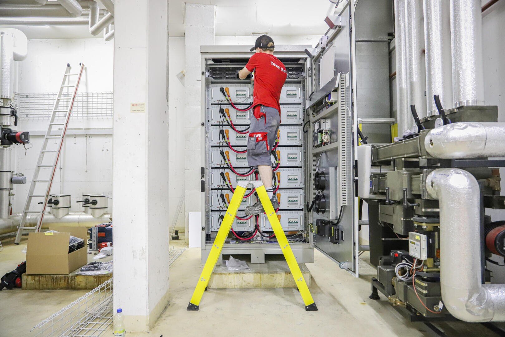 Ein Techniker in einem roten Hemd steht auf einer gelben Leiter und arbeitet an einer elektrischen Anlage in einem Industrieraum mit Rohren und Maschinen.
