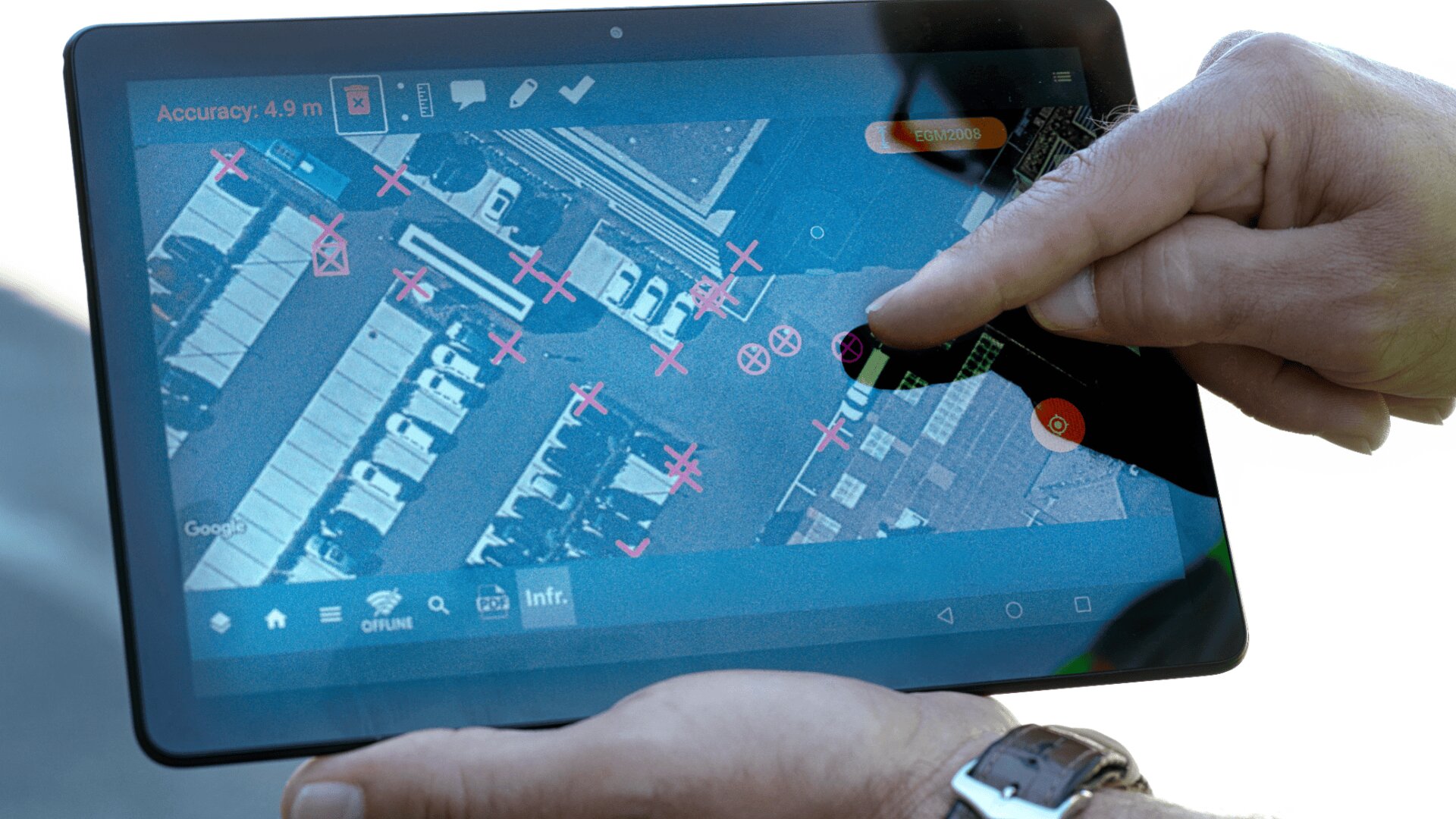 Eine Person benutzt ein Tablet, auf dem eine Satellitenkarte mit rosafarbenen X-Markierungen und Symbolen über geparkten Autos und Gebäuden angezeigt wird, und deutet auf einen bestimmten Ort auf dem Bildschirm.