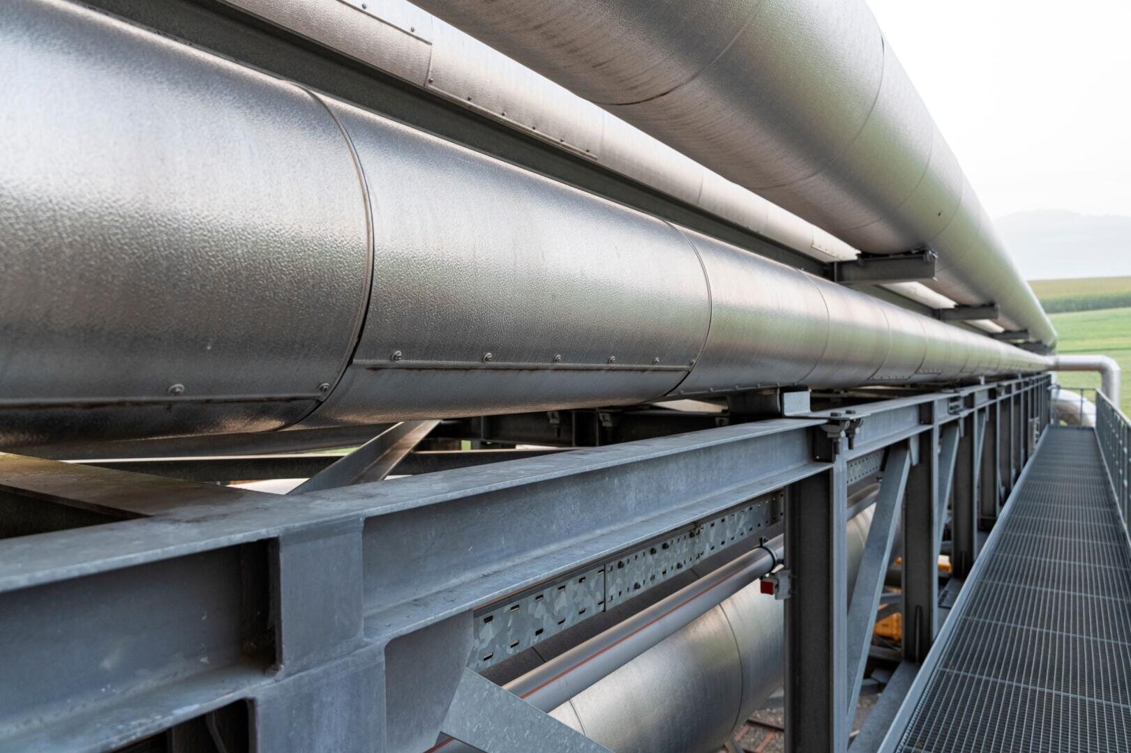 Große Industrierohre aus Metall verlaufen parallel zueinander auf einer Stahlkonstruktion, möglicherweise Teil eines Rohrleitungssystems, wobei im Hintergrund grüne Felder und der Himmel zu sehen sind.