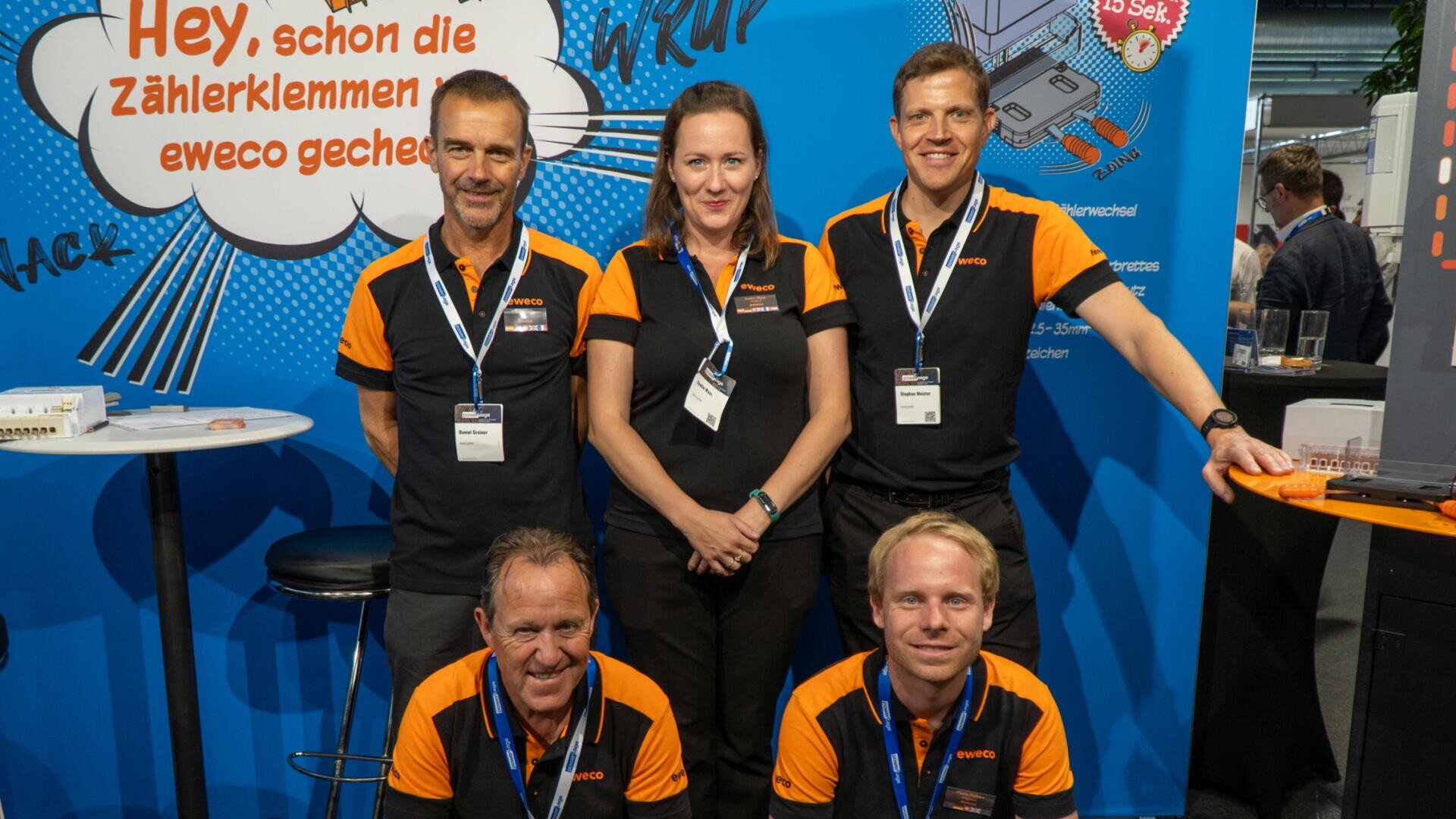 Fünf Personen in schwarzen und orangefarbenen Uniformen posieren und lächeln vor einem Messestand mit einem hellblauen Hintergrund, auf dem deutsche Texte und Illustrationen zum Thema Technik zu sehen sind.