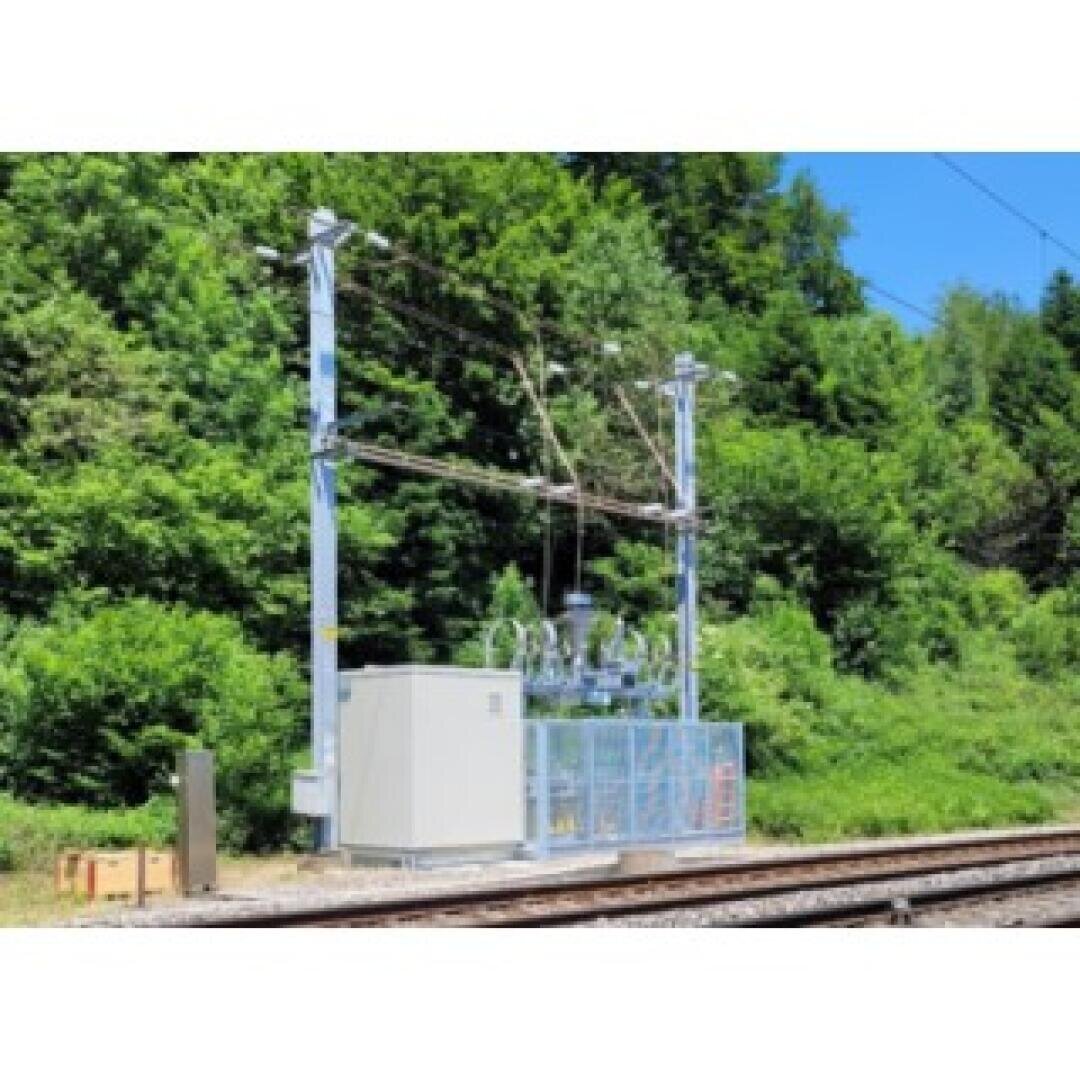 Ein kleines elektrisches Umspannwerk mit Transformatoren und Geräten befindet sich neben den Bahngleisen, umgeben von Grün und Bäumen, unter einem strahlend blauen Himmel. Die Oberleitungen sind mit dem Gebäude verbunden.