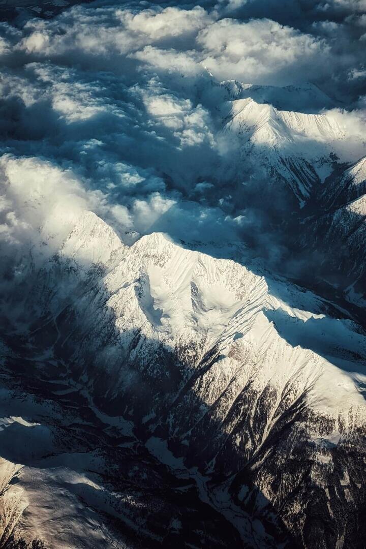 Aerial view of snow-covered mountain peaks partially shrouded by clouds, with sunlight casting dramatic shadows over the rugged landscape.