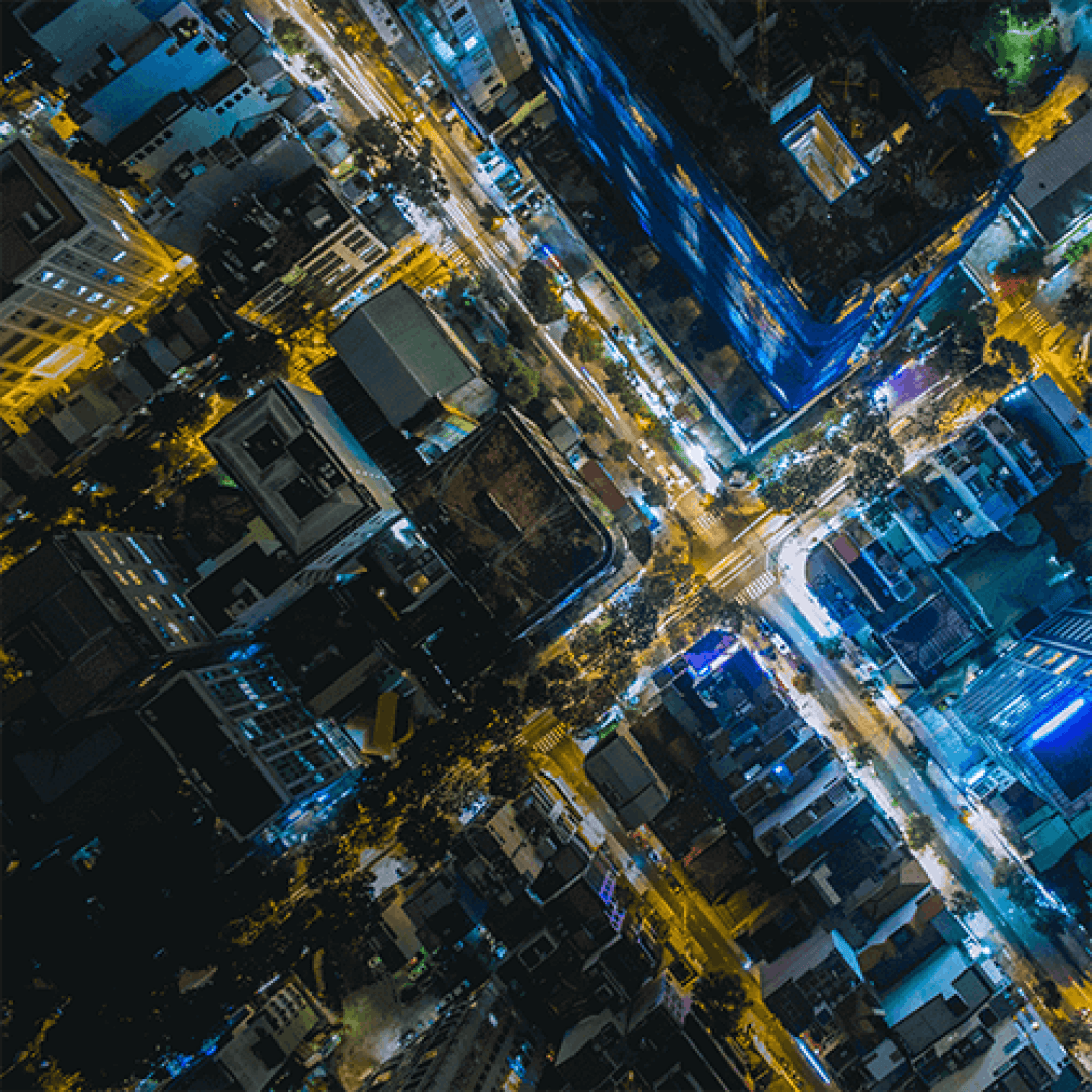 Luftaufnahme einer nächtlichen Stadtkreuzung mit beleuchteten Straßen, die ein X bilden, und Gebäuden, die mit gelben und blauen Lichtern beleuchtet sind.