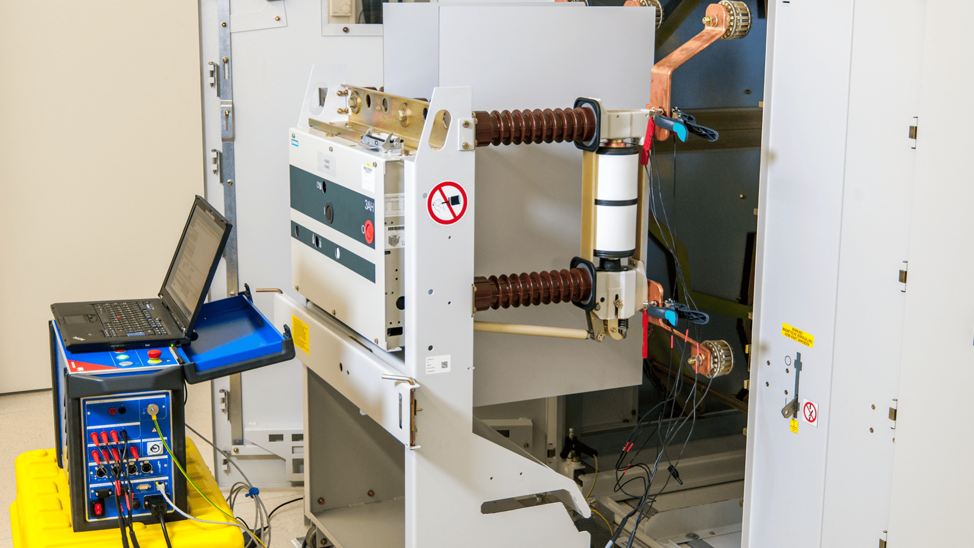 Eine Einrichtung zur Prüfung von Leistungsschaltern mit einem großen Leistungsschalter, der an Prüfgeräte und einen Laptop auf einem gelben Kasten angeschlossen ist, in einem Technikraum mit Schalttafeln.