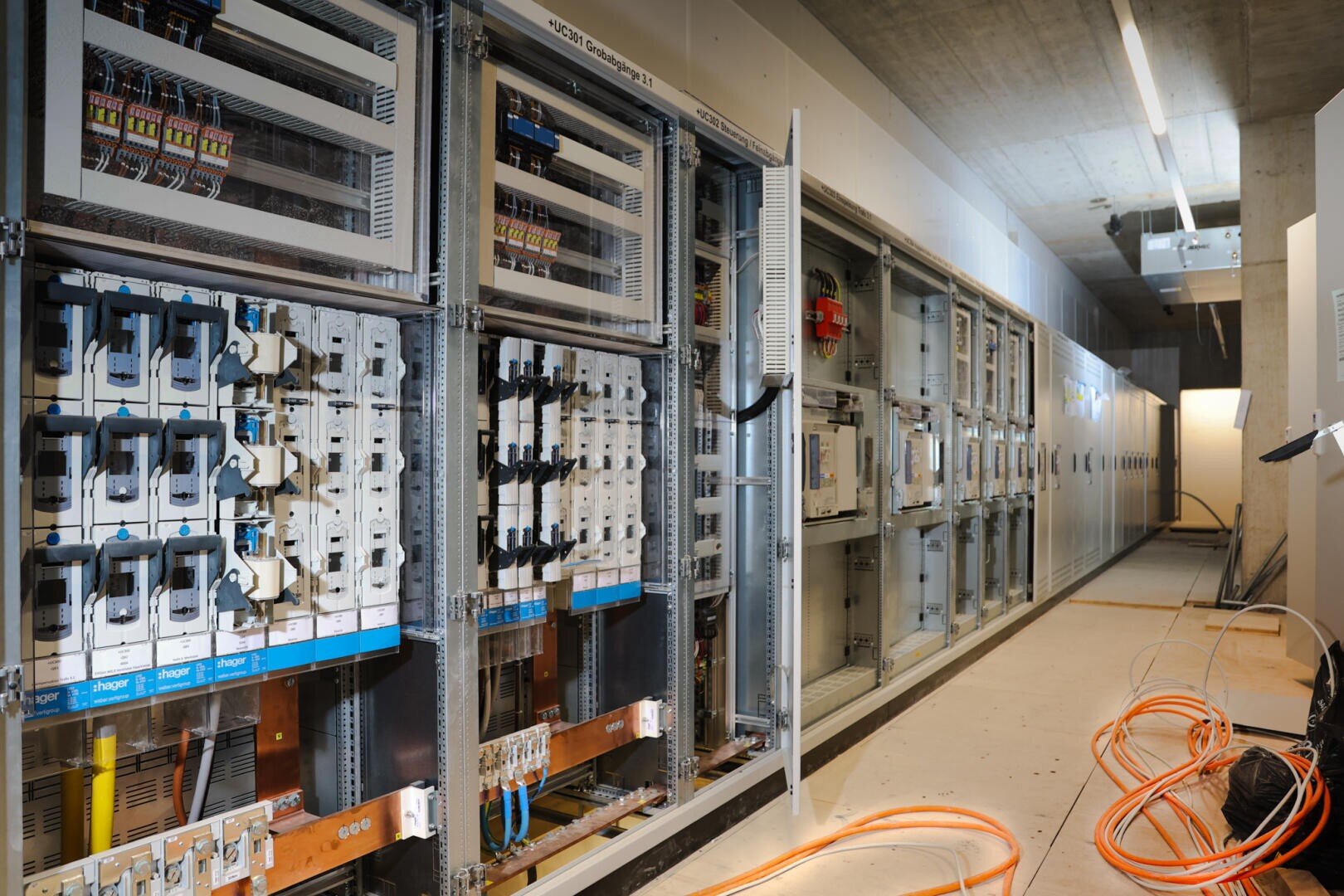 Reihen von elektrischen Schalttafeln und Leistungsschaltern in einem im Bau befindlichen Industrieraum mit freiliegenden Leitungen und orangefarbenen Kabeln auf dem Boden. Überkopf-Leuchtstoffröhren erhellen den unvollendeten Raum.