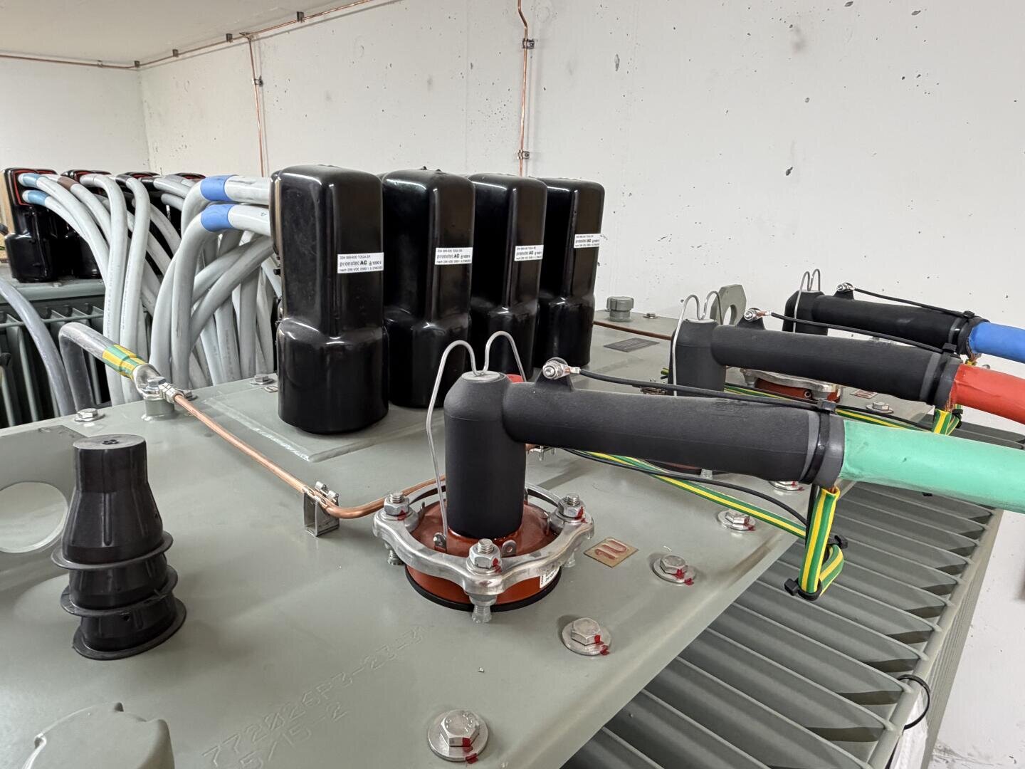 Nahaufnahme von elektrischen Komponenten, die an einem Transformator montiert sind, mit isolierten Kabeln, Steckern und Erdungsdrähten, die an der Metalloberfläche in einer Industrieumgebung befestigt sind.