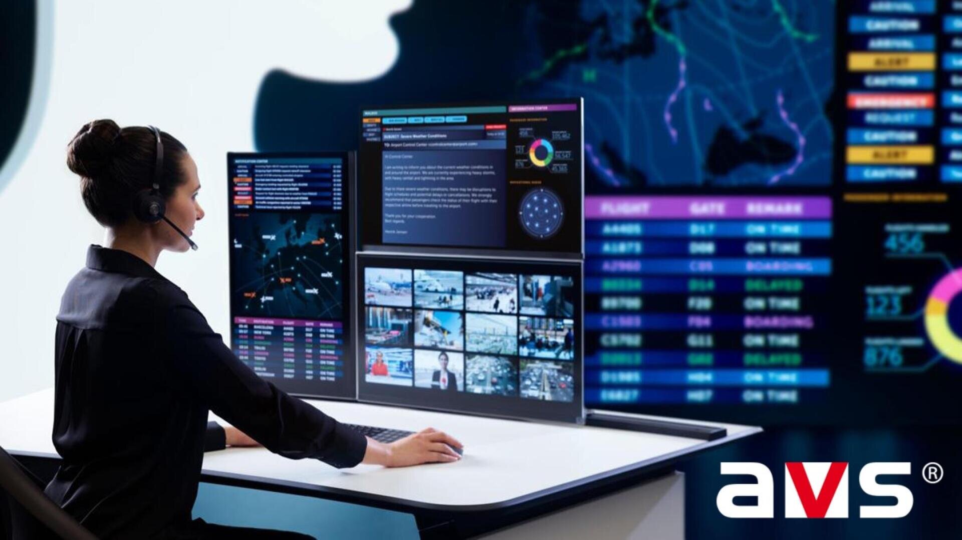 Eine Frau mit einem Headset sitzt in einem Hightech-Kontrollraum an einem Schreibtisch mit mehreren Monitoren, auf denen Karten, Diagramme und Überwachungsmaterial angezeigt werden. In der rechten unteren Ecke ist das AVS-Logo zu sehen.