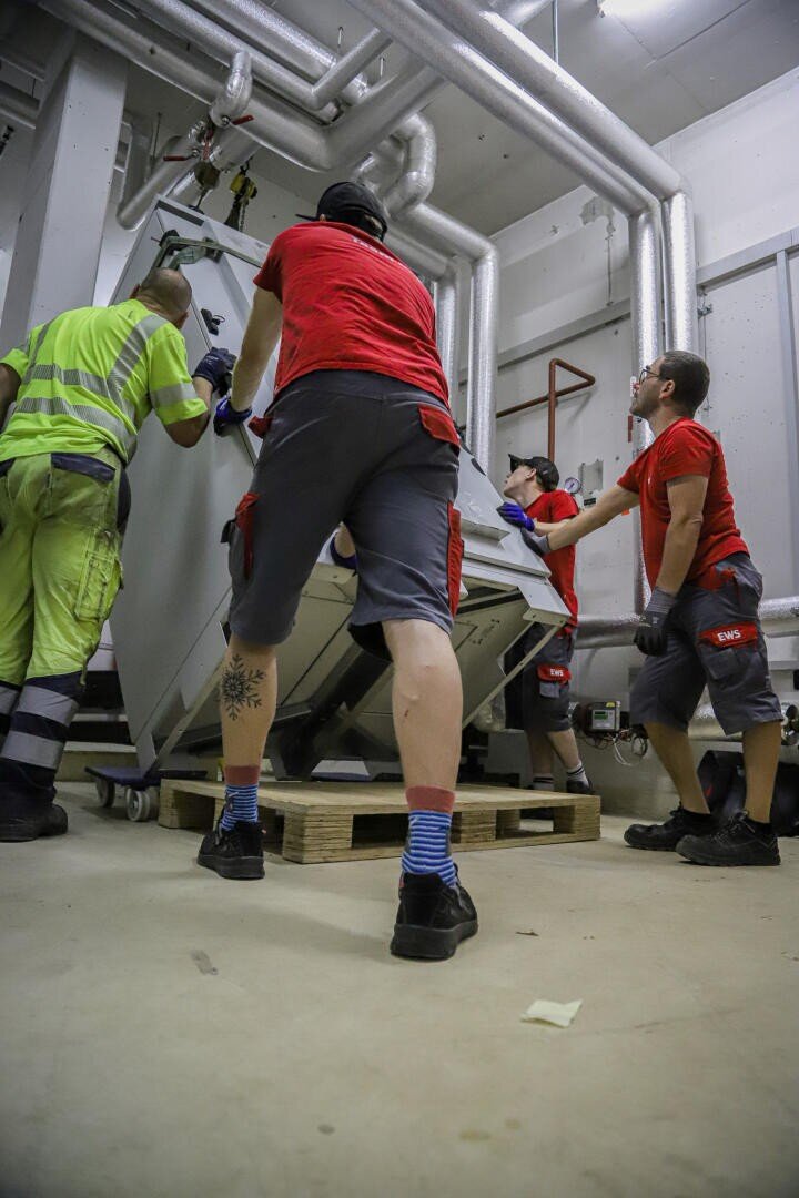 Vier Arbeiter, drei in roten Hemden und einer in einem gelb-grünen Outfit, bewegen eine große, schwere Maschine auf einer Palette in einem Industrieraum mit freiliegenden Rohren und einem Betonboden.