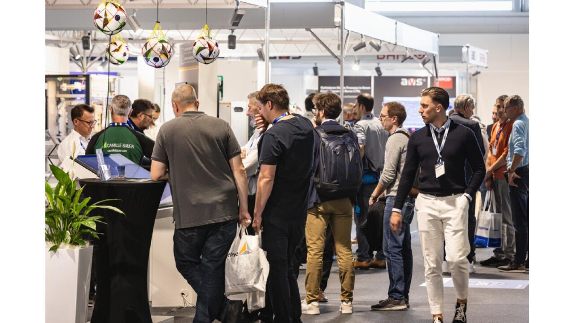 A group of people attend a busy trade show or exhibition, standing and walking between booths displaying products and banners. Some attendees carry bags and wear lanyards with name badges.