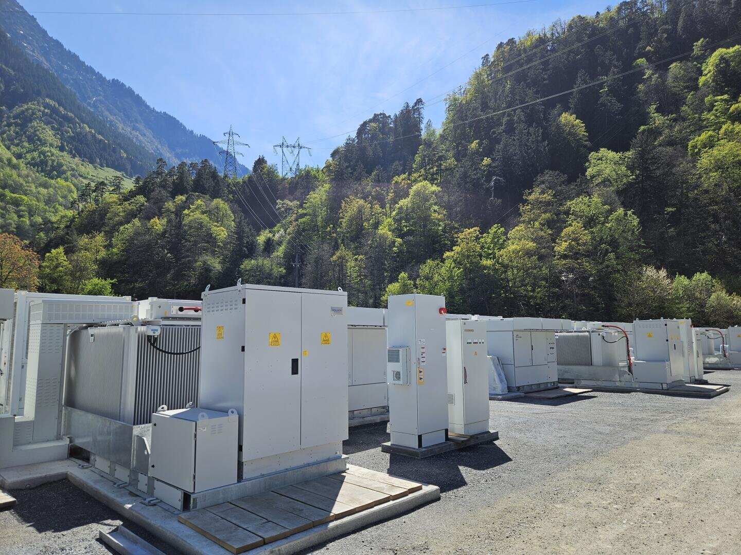 Reihen von weißen Elektrogeräten und Lagereinheiten stehen im Freien auf einer Kiesfläche, umgeben von grünen, bewaldeten Bergen unter einem klaren blauen Himmel mit sichtbaren Stromleitungen darüber.