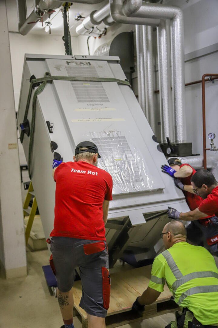 Drei Arbeiter in Schutzkleidung führen eine große, gekippte Industriemaschine auf eine Holzplattform in einem Maschinenraum mit freiliegenden Rohren. Zwei tragen rote Hemden mit der Aufschrift "Team Rot", einer trägt ein gelbes Hemd mit hoher Sichtbarkeit.