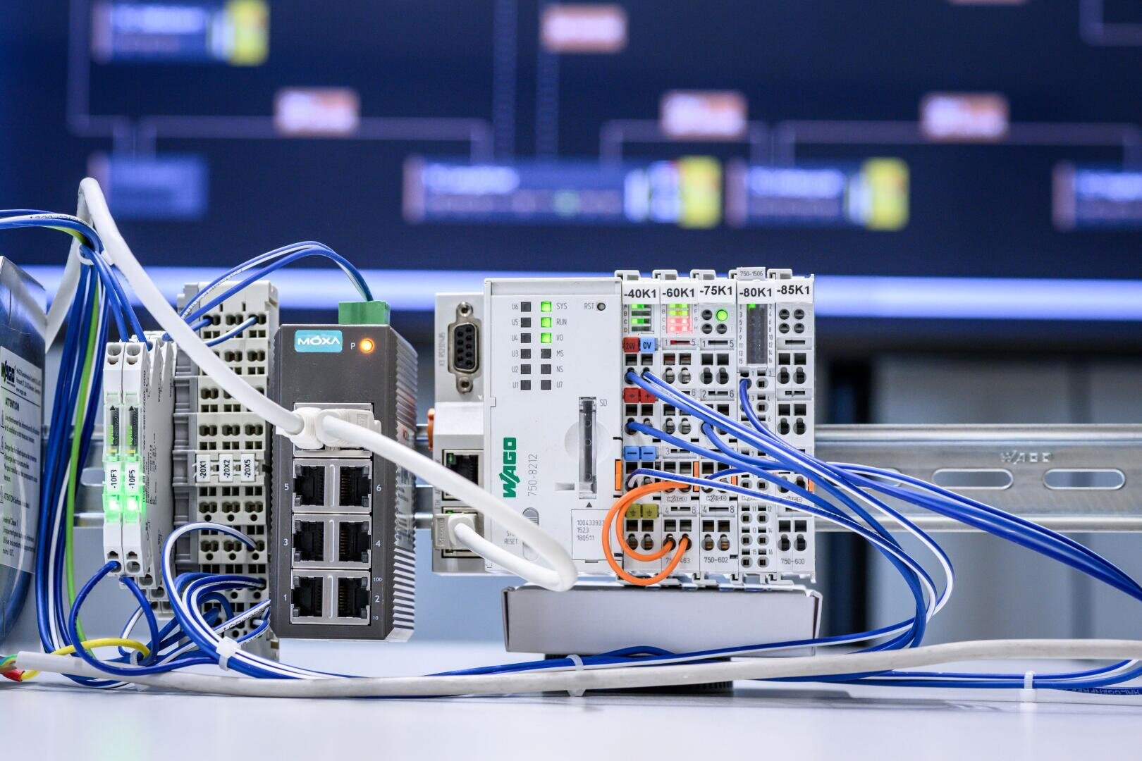 Nahaufnahme einer industriellen Automatisierungseinrichtung mit Ethernet-Kabeln, einem Netzwerk-Switch und auf einer Schiene montierten SPS-Modulen, mit einer unscharfen Steuerungsschnittstelle im Hintergrund.