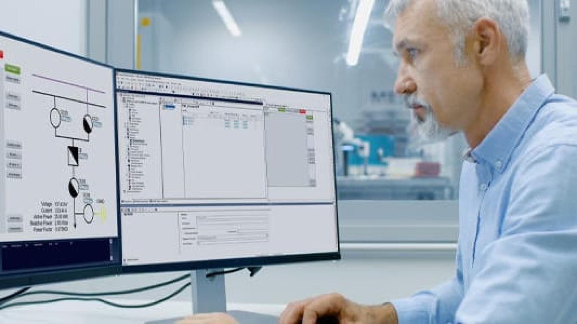 Ein Mann mit grauem Haar und Bart arbeitet an einem Schreibtisch und schaut aufmerksam auf zwei Computermonitore, auf denen technische Diagramme und Software-Schnittstellen in einer modernen Büro- oder Laborumgebung angezeigt werden.