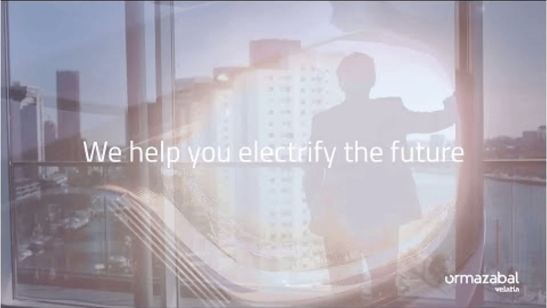A person stands by a large window overlooking city buildings. Overlaid text reads, We help you electrify the future. The Urmazabal Velatia logo appears in the bottom right corner.