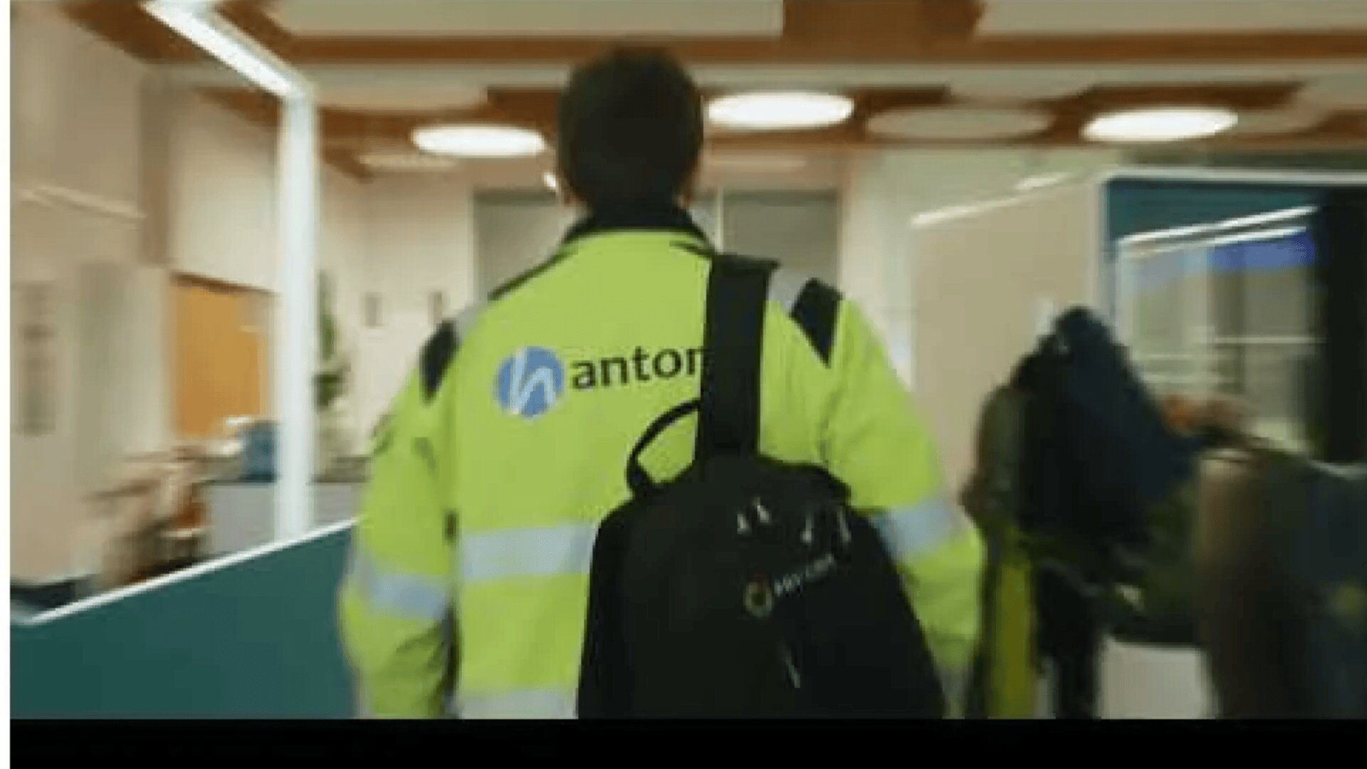 Person in einer Warnweste mit Nanto und einem blauen Logo auf dem Rücken, die einen schwarzen Rucksack trägt und in einem modernen, gut beleuchteten Büro oder Flur geht.