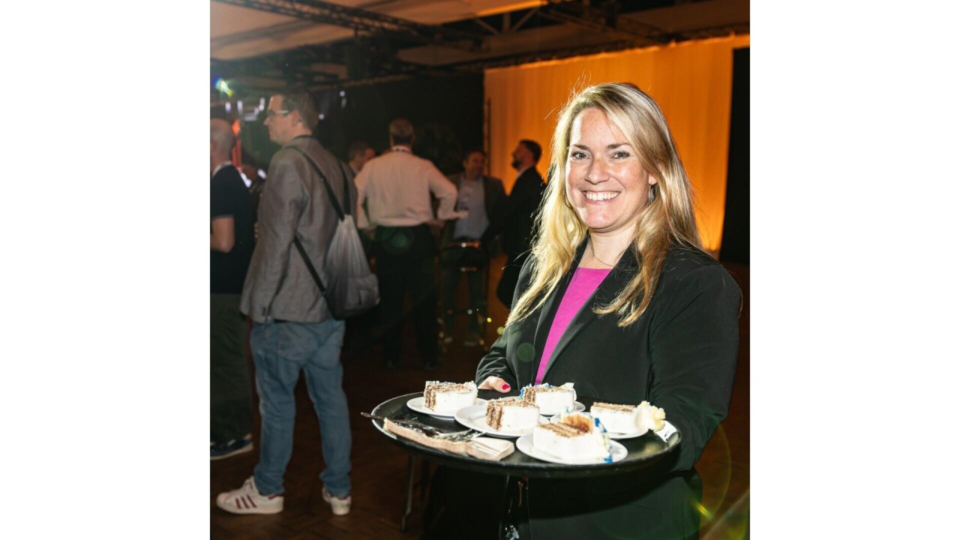 A smiling woman in a black blazer holds a tray with slices of cake at an indoor event. Other people are visible in the background, and the room is warmly lit.