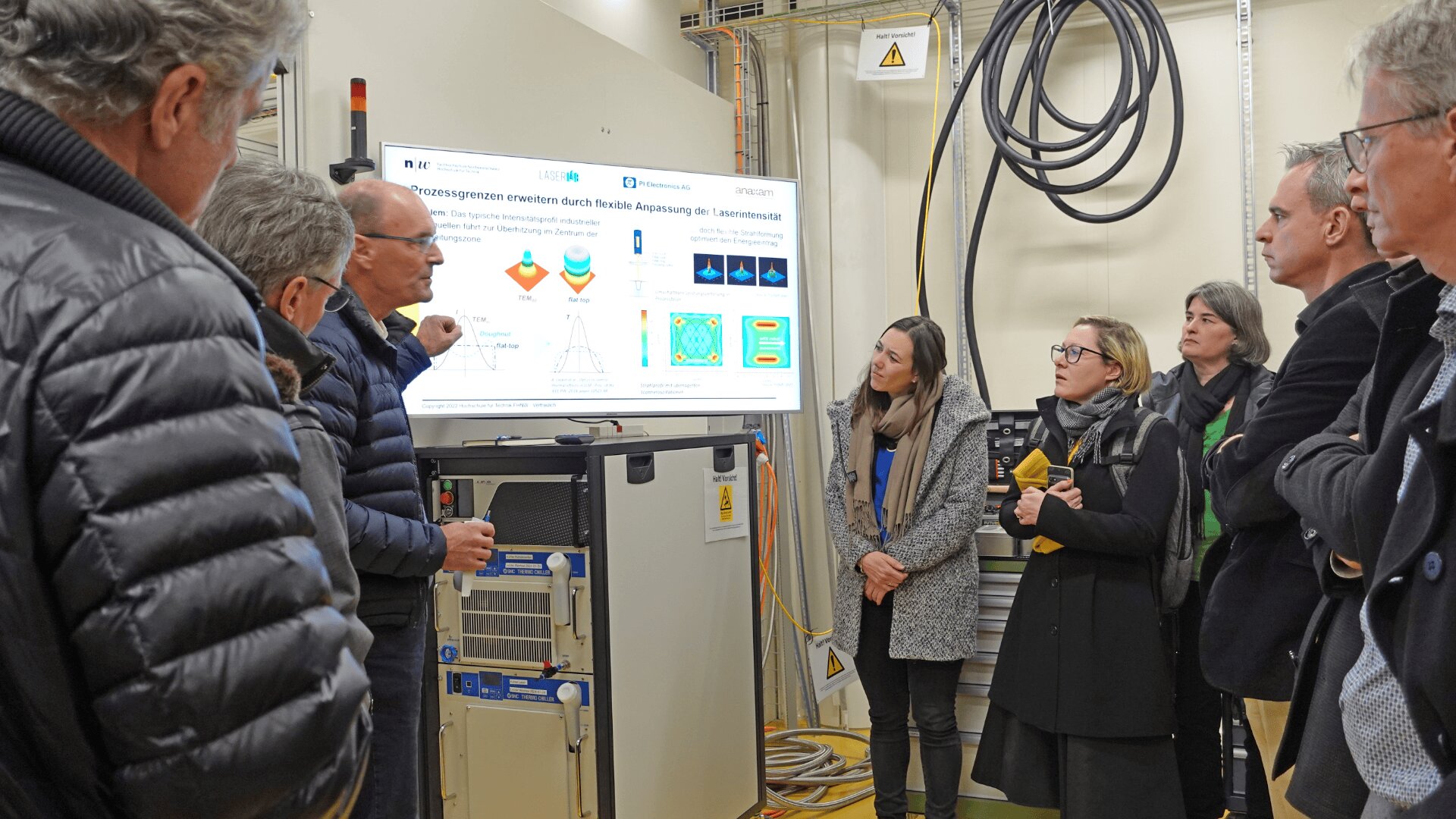 Eine Gruppe von Personen steht in einem Labor um einen Moderator herum, hört zu und schaut auf einen großen Bildschirm, auf dem wissenschaftliche Diagramme und deutscher Text zu sehen sind. Im Hintergrund sind Laborgeräte und Kabel zu sehen.