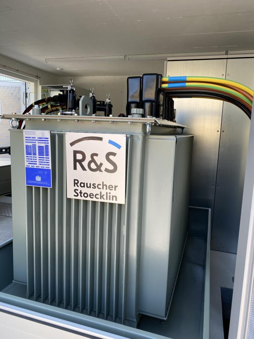 Ein großer grauer elektrischer Transformator mit der Aufschrift R&S Rauscher Stoecklin ist in einem Innenraum installiert. An der Oberseite sind farbige Kabel angeschlossen, und am Gerät ist eine technische Informationstafel angebracht.