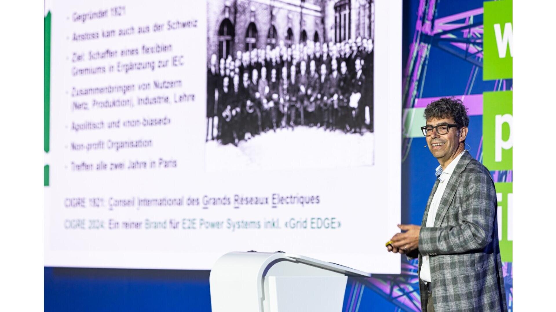 A man in glasses and a suit jacket stands smiling at a podium, giving a presentation with a projected slide behind him featuring text in German and a black-and-white historical group photo.