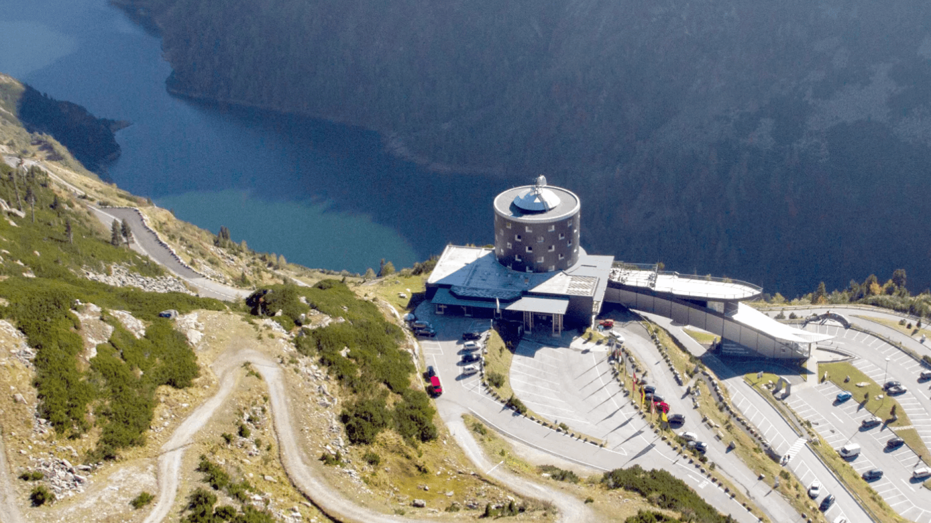 Luftaufnahme eines modernen, runden Gebäudes mit Kuppeldach und kreisrunden Fenstern, umgeben von Parkplätzen und kurvenreichen Straßen in einer bergigen Gegend in der Nähe eines Sees.