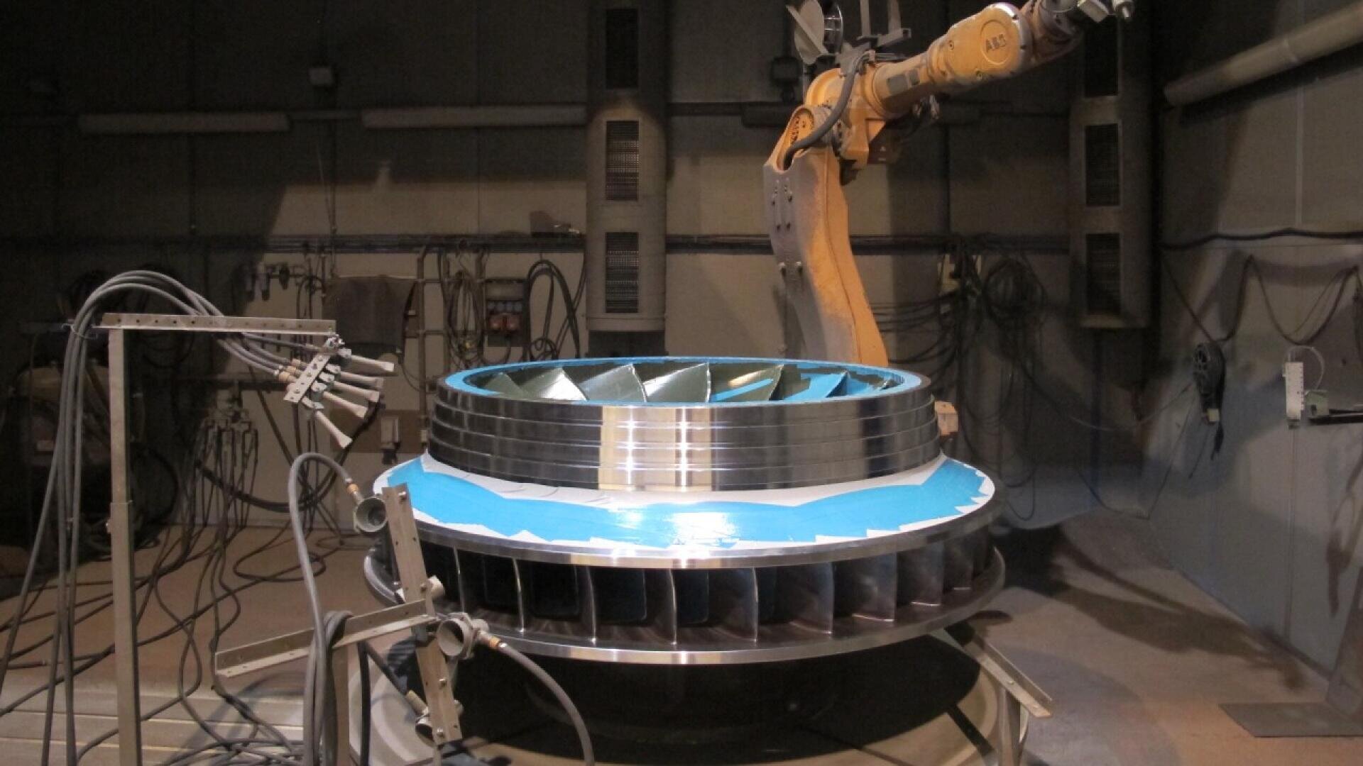A robotic arm works on a large, metallic turbine component inside an industrial facility, surrounded by cables, pipes, and various equipment. The turbine has blue tape applied to its surface.