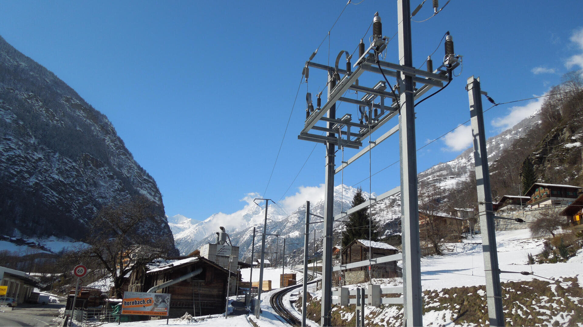 A snow-covered village in a mountain valley with wooden houses, power lines, electricity pylons and a bright blue sky. Rocky, wooded mountains rise up on either side and a snow-covered road winds its way through the scenery.