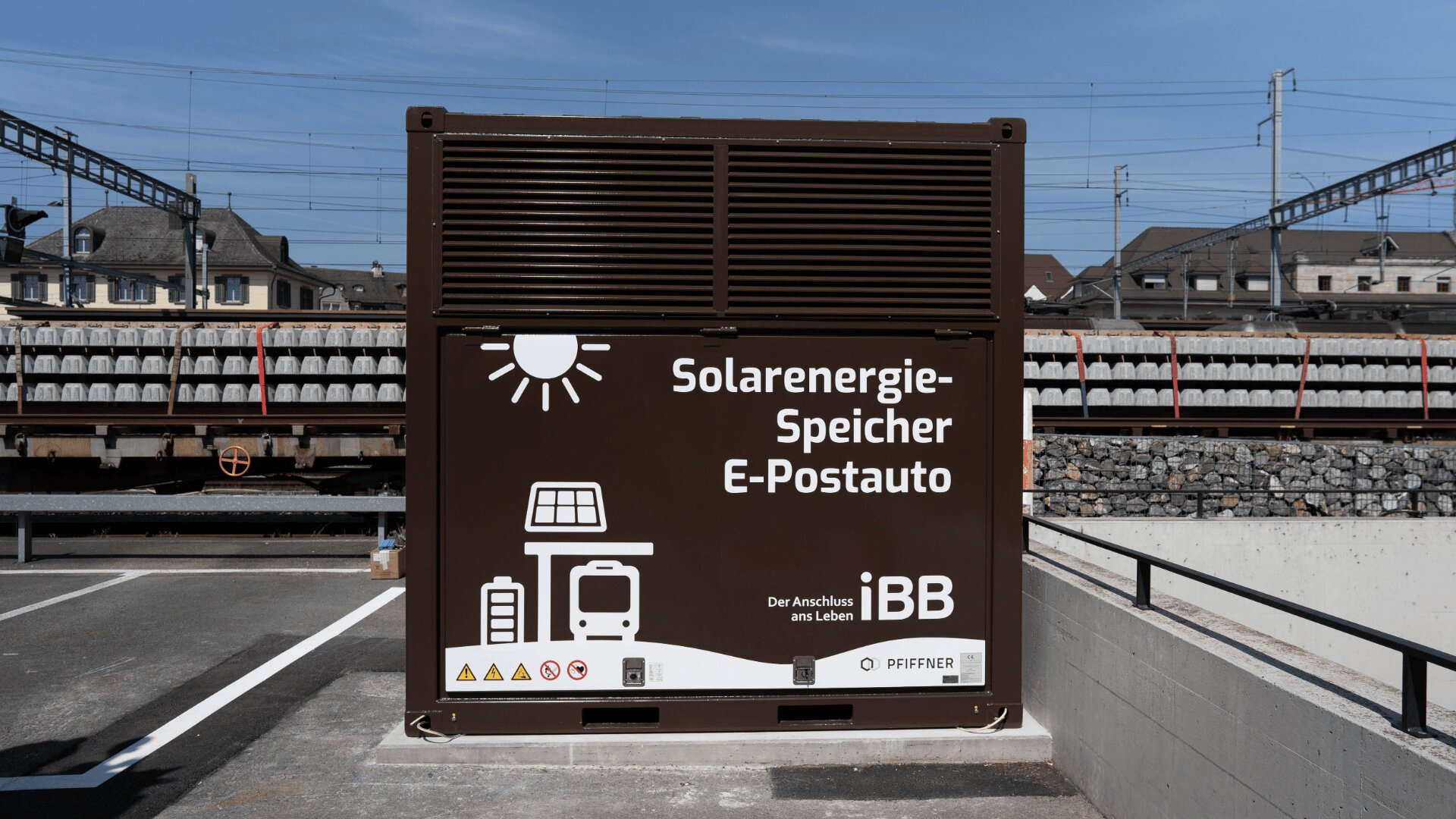 Ein großer brauner Solarenergiespeicher mit der Aufschrift Solarenergie-Speicher E-Postauto steht auf einem Parkplatz in der Nähe der Bahngleise. Auf seiner Vorderseite sind die Symbole einer Sonne, eines Busses und einer Ladestation abgebildet.