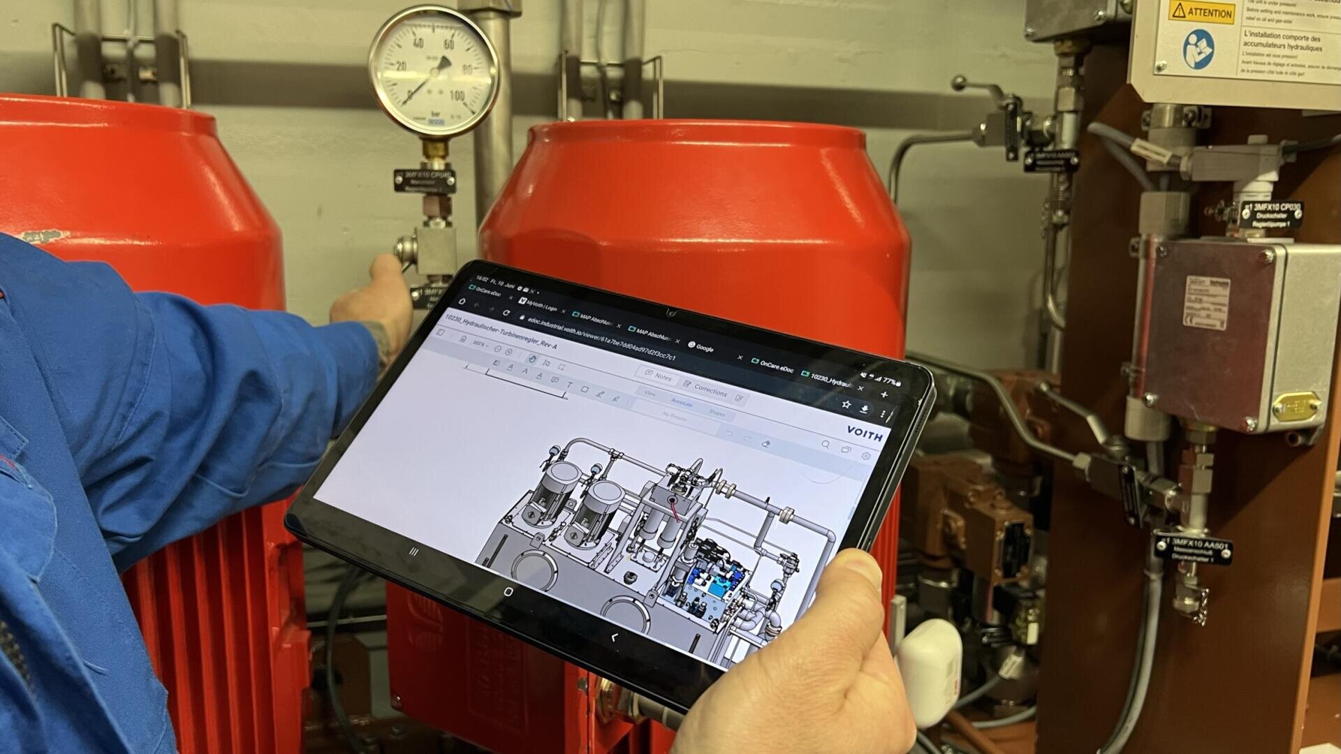 Eine Person in Blau hält ein Tablet, auf dem ein industrielles Schema abgebildet ist, vor eine rote Maschine und ein Messgerät und scheint das digitale Diagramm mit der physischen Ausrüstung in einer technischen Umgebung zu vergleichen.