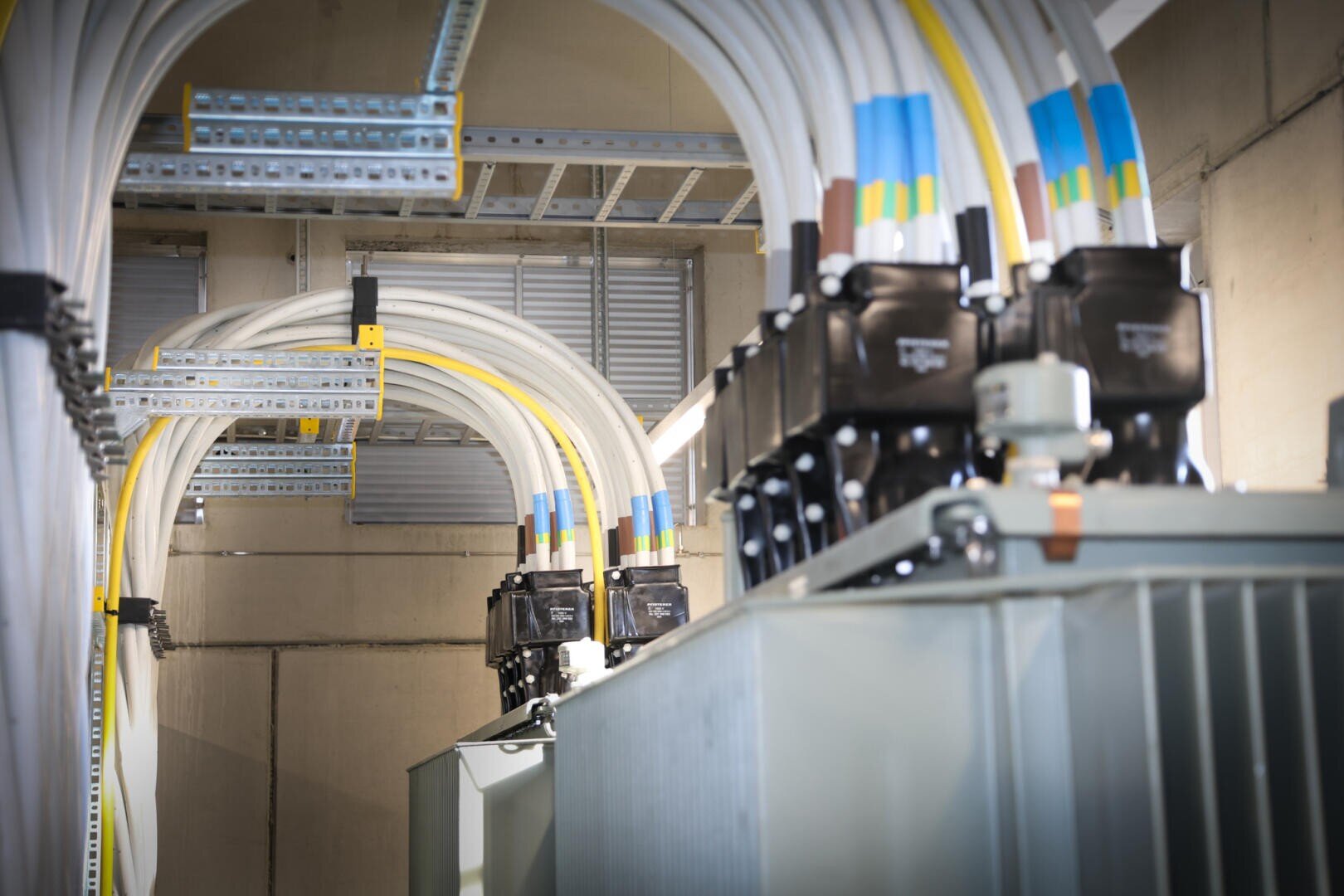 Große elektrische Kabel und Steckverbinder sind an Industrietransformatoren in einem Versorgungsraum befestigt, mit Kabeltrassen über dem Kopf und Betonwänden im Hintergrund.