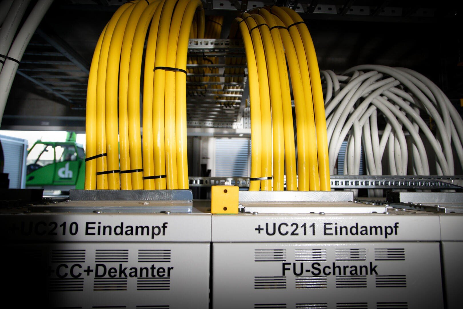 Gelbe und weiße Netzwerkkabel sind geordnet und an beschriftete Serverschränke angeschlossen, auf deren Metalloberflächen Etiketten wie +UC210 Eindampf und +UC211 Eindampf zu sehen sind.