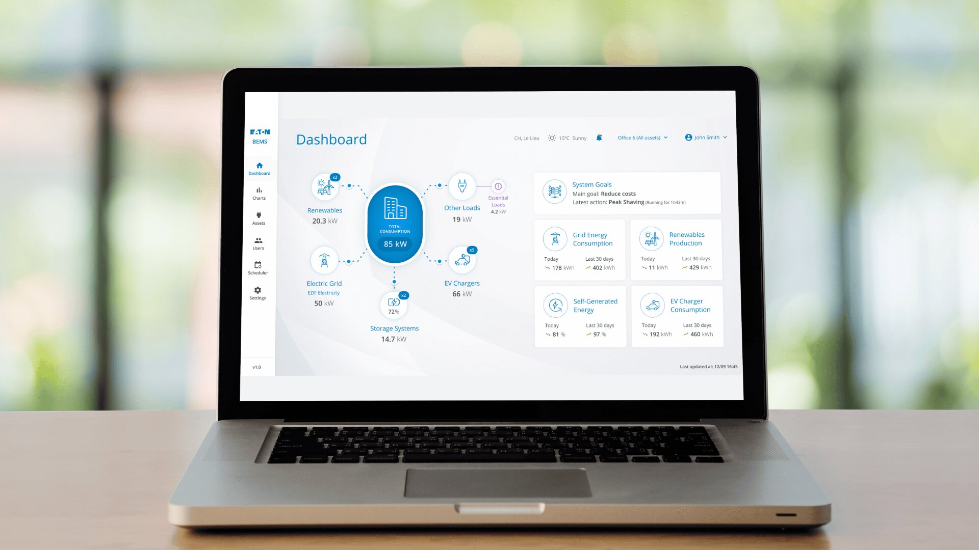 Ein Laptop auf einem Holztisch zeigt ein Armaturenbrett mit Diagrammen und Daten über erneuerbare Energien, das Stromnetz, Ladestationen für Elektrofahrzeuge und Speichersysteme. Der Hintergrund ist mit Grün- und Brauntönen verschwommen.