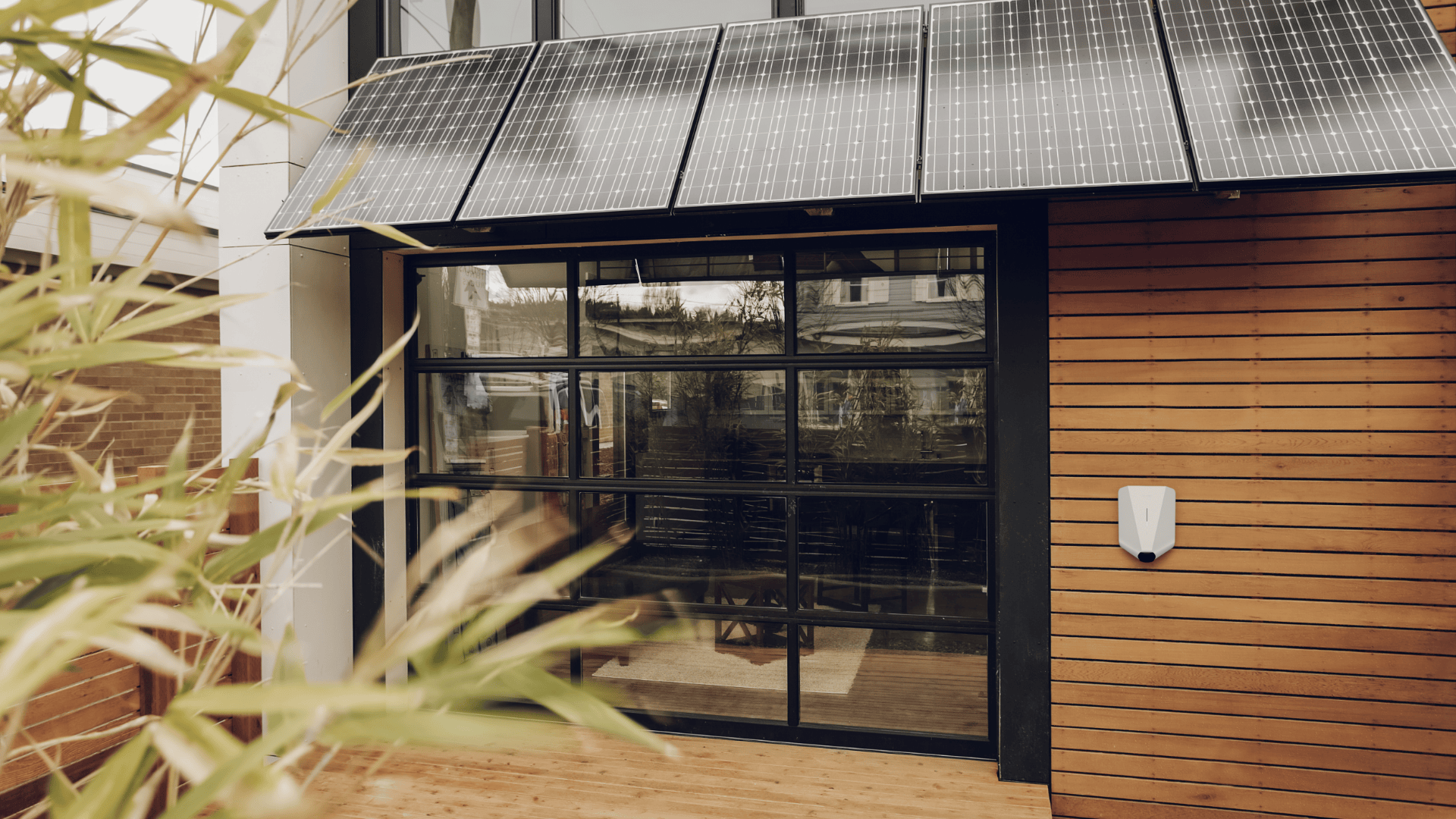 Ein modernes Gebäude mit Holzverkleidung, großen Garagentoren aus Glas, Sonnenkollektoren auf dem Dach und einer Ladestation für Elektrofahrzeuge an der Wand. Im Vordergrund ist grünes Laub zu sehen.