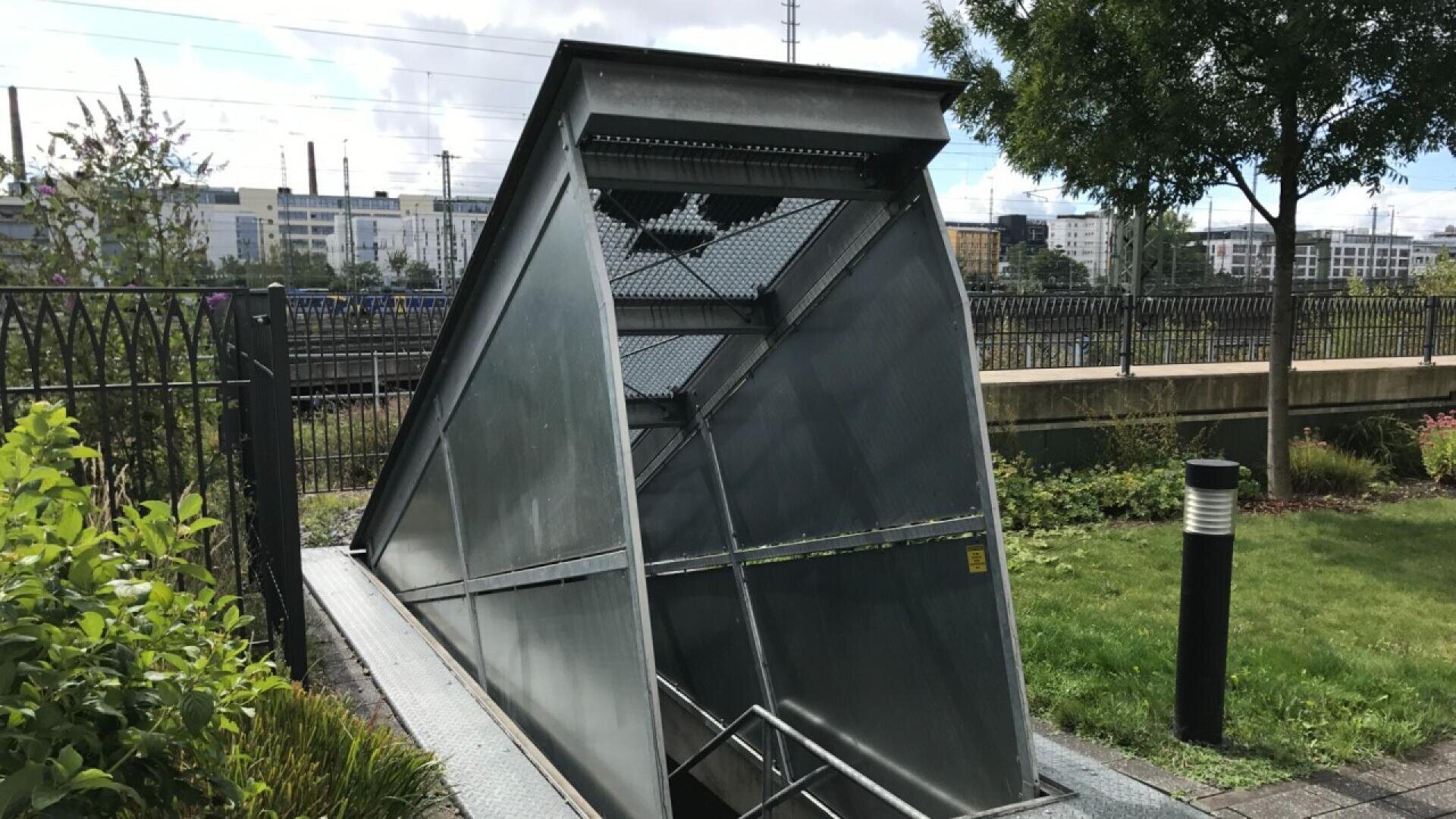 A modern, angular metal entrance leads underground beside a grassy area and garden, near railroad tracks and buildings in the background. The structure features sharp lines and a stairway descending below ground.